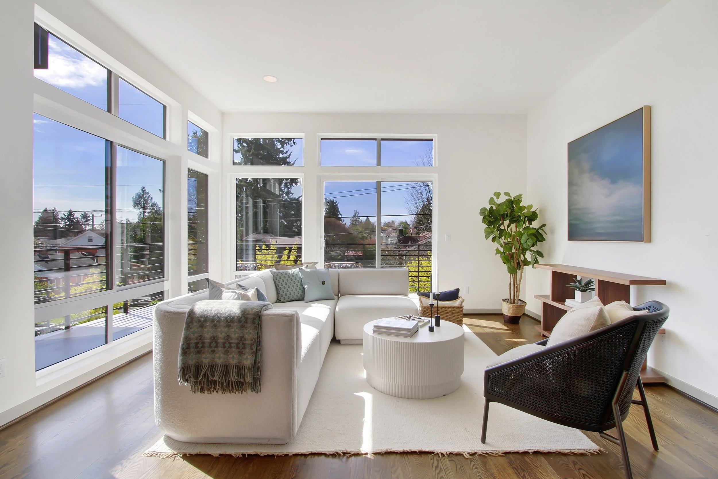 Bright living room with white walls and large windows showing a sunny sky, trees, and neighboring houses. Contains a white sectional sofa with pillows, a round white coffee table with books and decor, a black armchair, a wicker basket, a large plant, and wall art. Hardwood floors and a cozy, modern decor style.