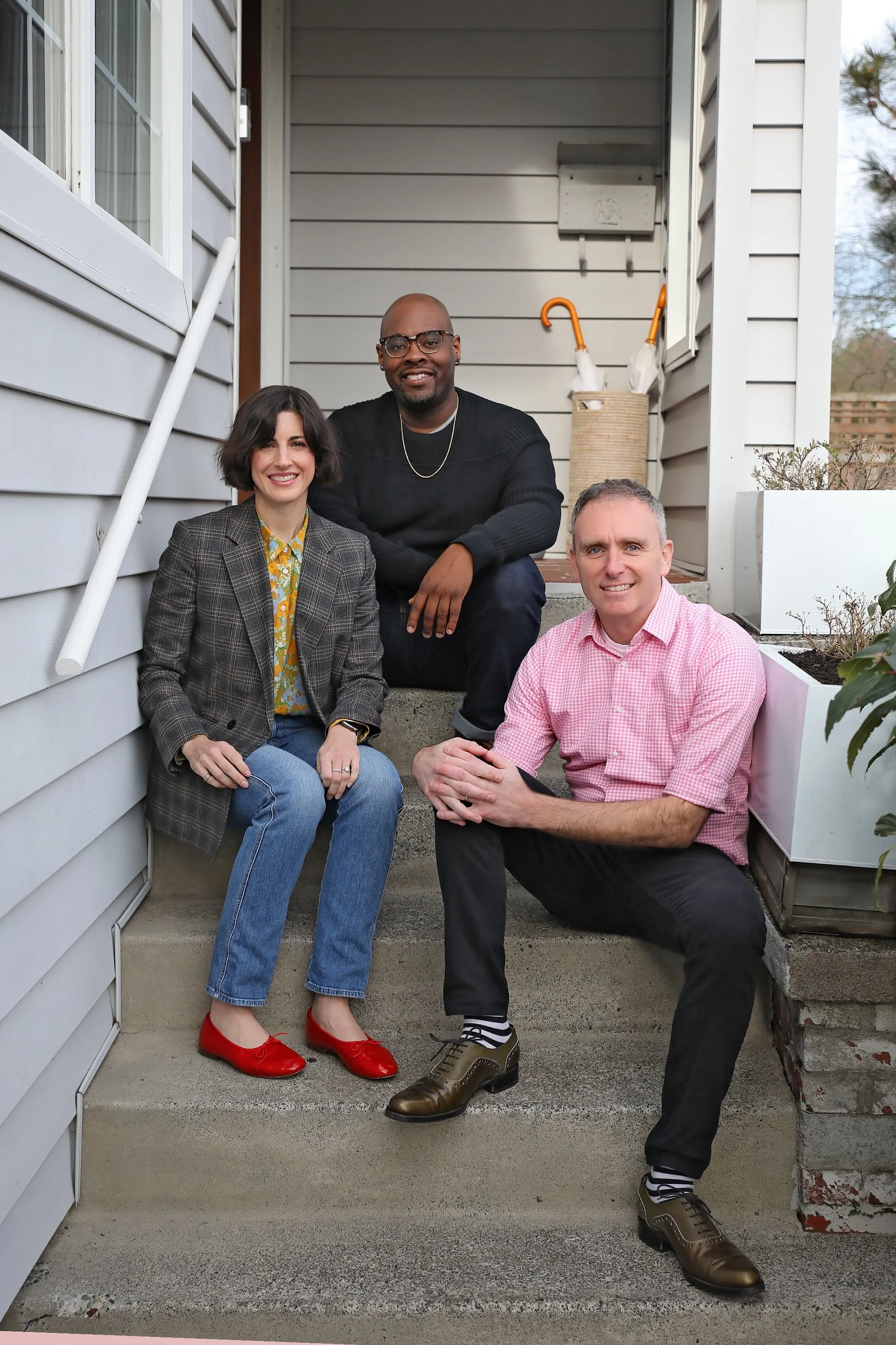 A group of three people sitting on the front steps of a house, smiling at the camera. The woman on the left wears a plaid blazer, floral blouse, blue jeans, and red shoes. The man in the middle wears black glasses, a black sweater, and dark pants. The man on the right wears a pink checked shirt and black pants.