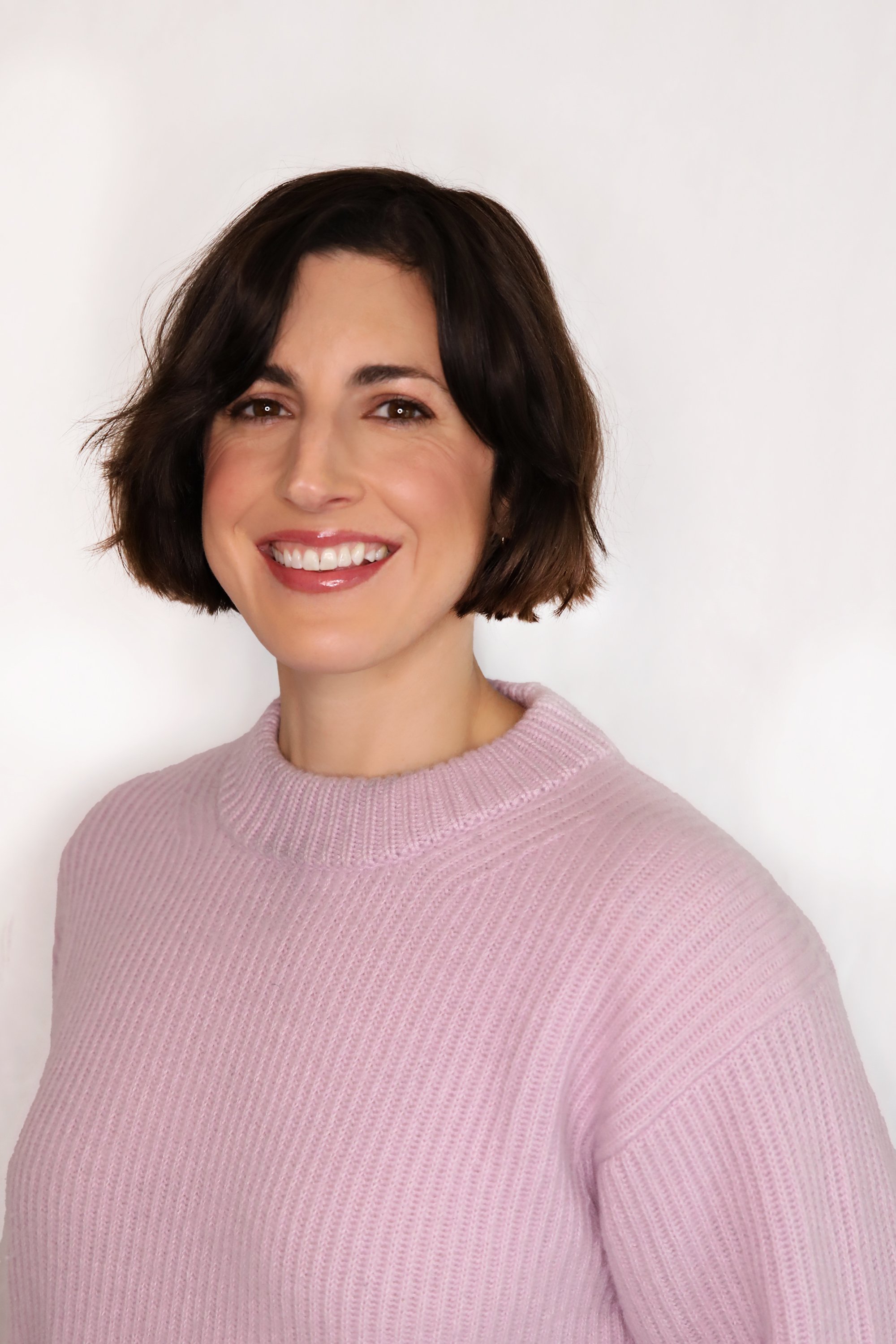 Portrait of a woman with short brown hair, smiling, wearing a light purple sweater, standing against a plain white background.