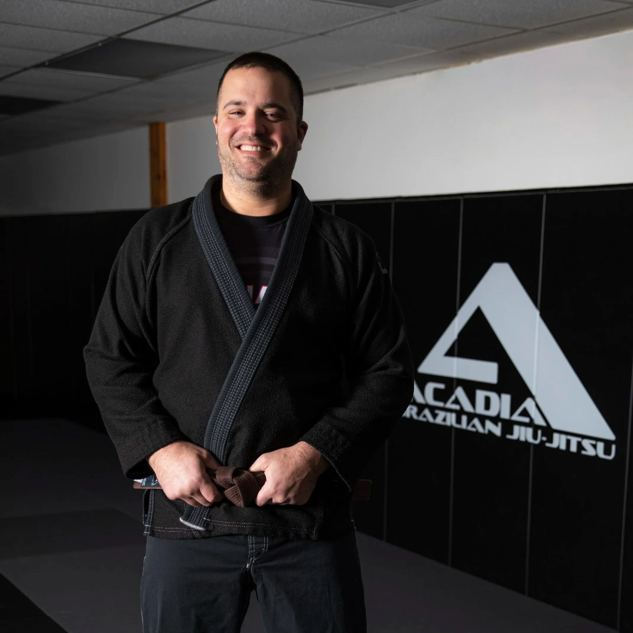 Man standing inside Brazilian Jiu-Jitsu gym, smiling, wearing a black gi with a brown belt, in front of a wall with the logo 'Acadía Brazilian Jiu-Jitsu'.