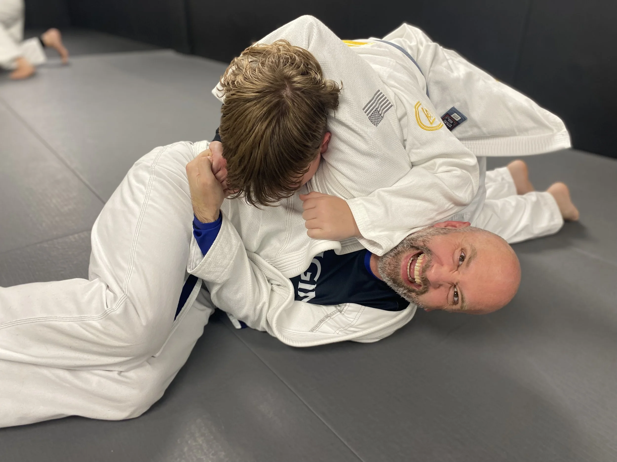 Two men practicing Brazilian Jiu-Jitsu on a gray mat, one in a white gi and the other in a dark gi, engaged in a grappling move.