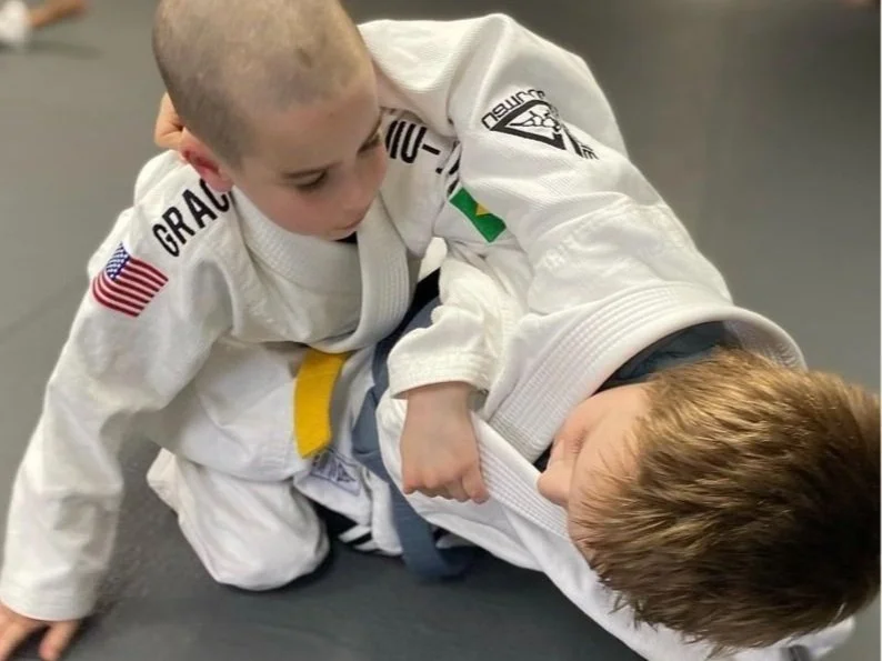 Two young children practicing Brazilian Jiu-Jitsu on a mat, with one child on top in a dominant position and the other child underneath, both wearing gis.