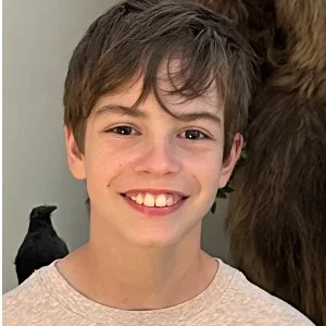 Two children stand in front of a penguin exhibit at a zoo, with penguins and a rocky background visible behind them.