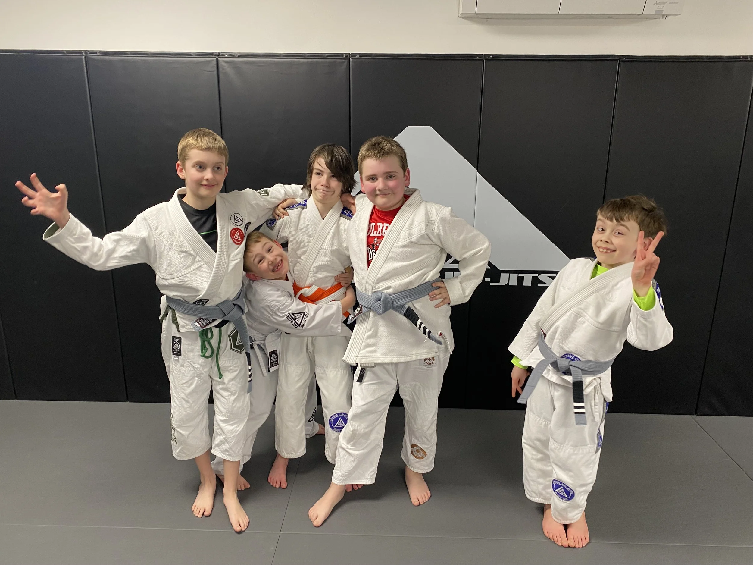 Group of five young boys in Brazilian Jiu-Jitsu uniforms standing on a mat in a gym, smiling, posing, and making gestures after practice or competition.