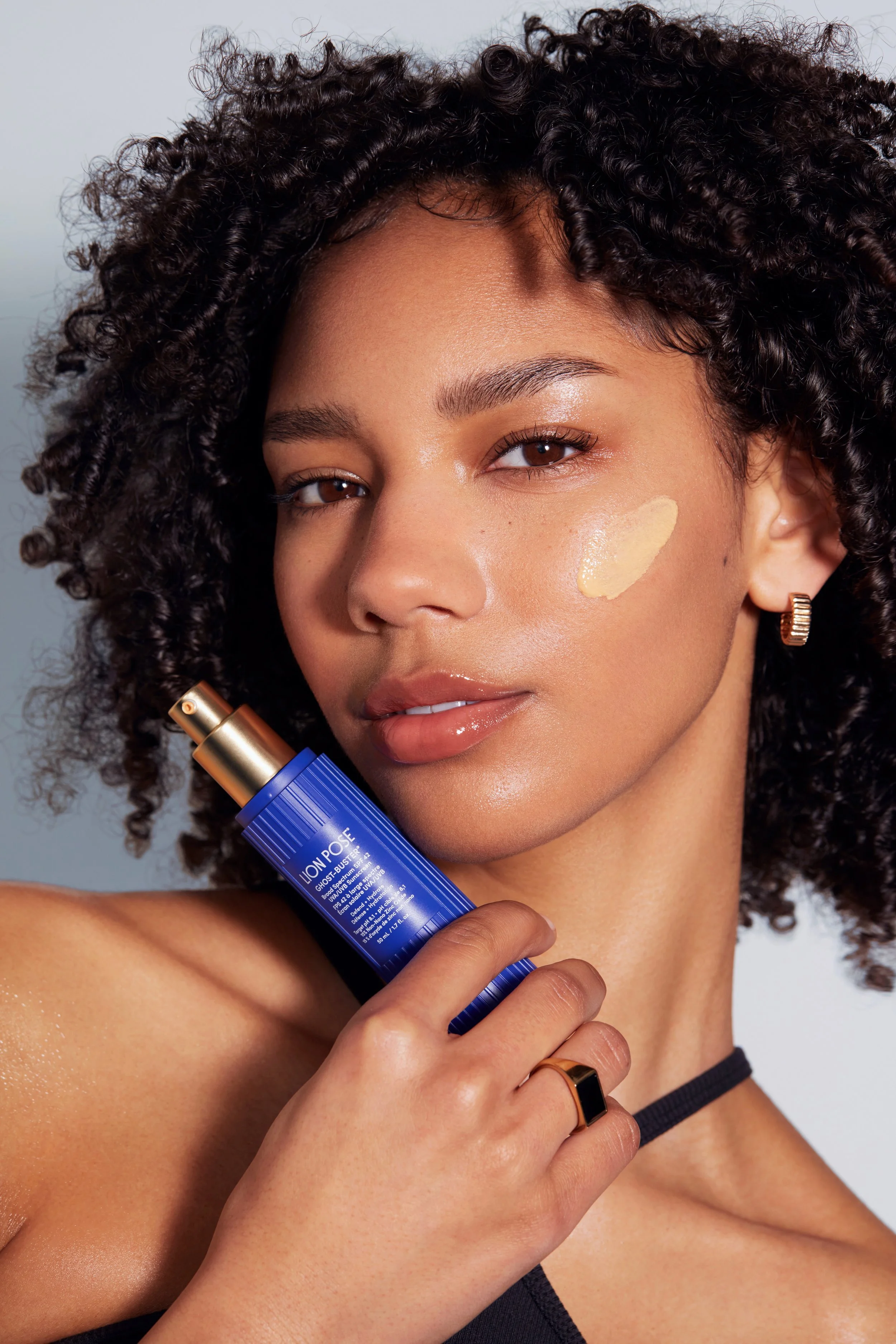 A woman with curly hair applying a skincare product from a blue bottle labeled LIOVEL PORE GLOSS BUST to her face.