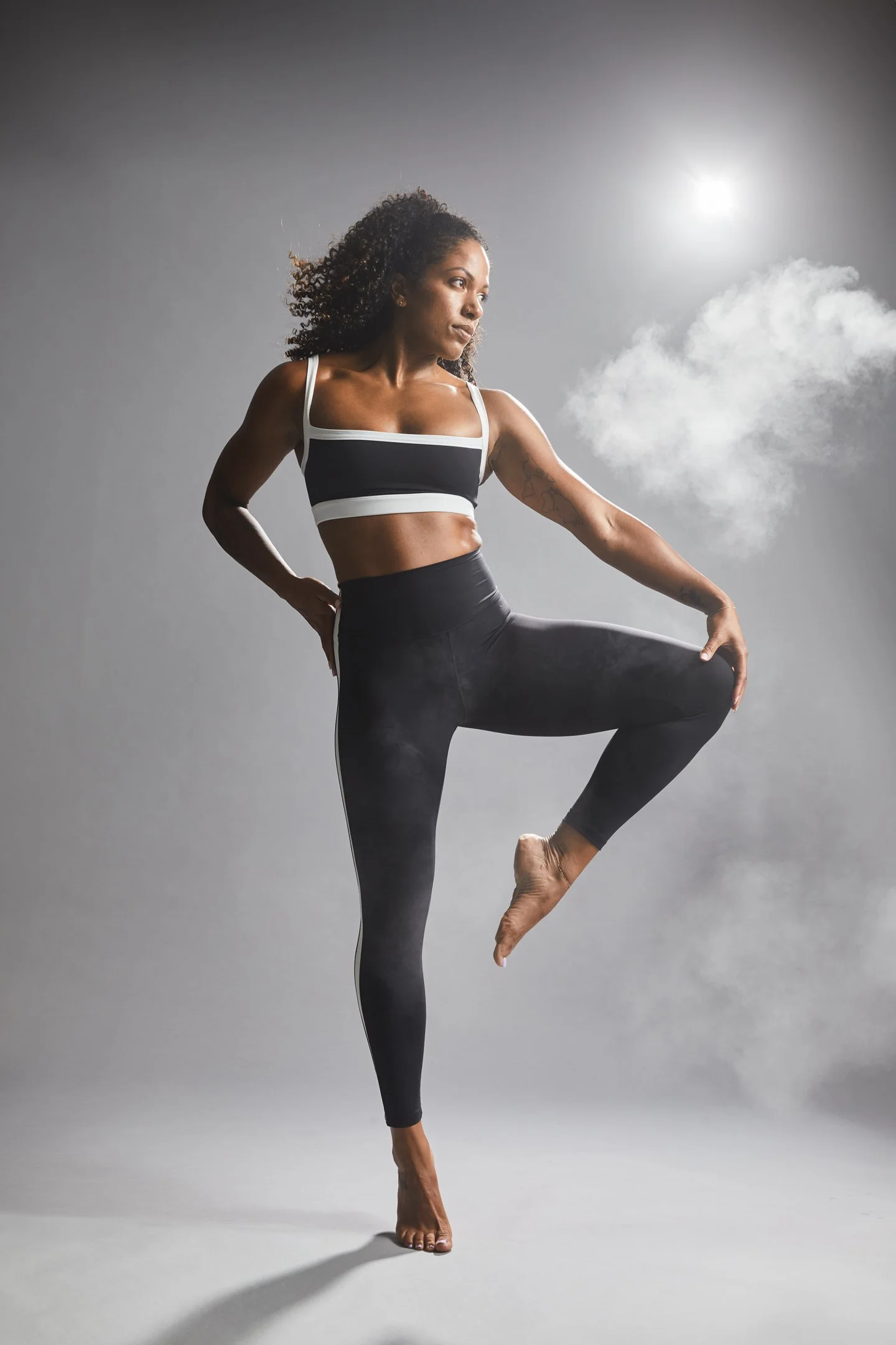 A woman practicing yoga outdoors at sunrise, standing on one foot with her other leg bent and foot resting on her thigh, wearing a black and white sports bra and high-waisted black leggings.