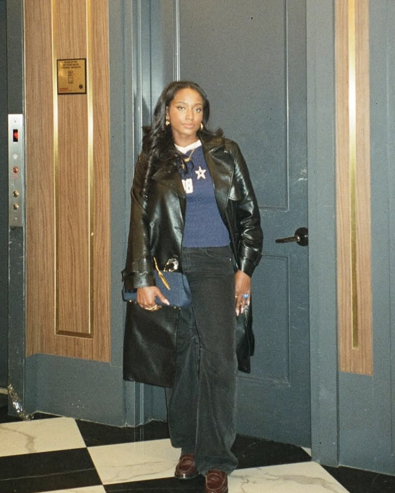 A woman with long, wavy black hair standing in an elevator lobby, wearing a black leather jacket, a blue sweater with stars, black pants, and brown shoes, holding a blue purse and a black bag.
