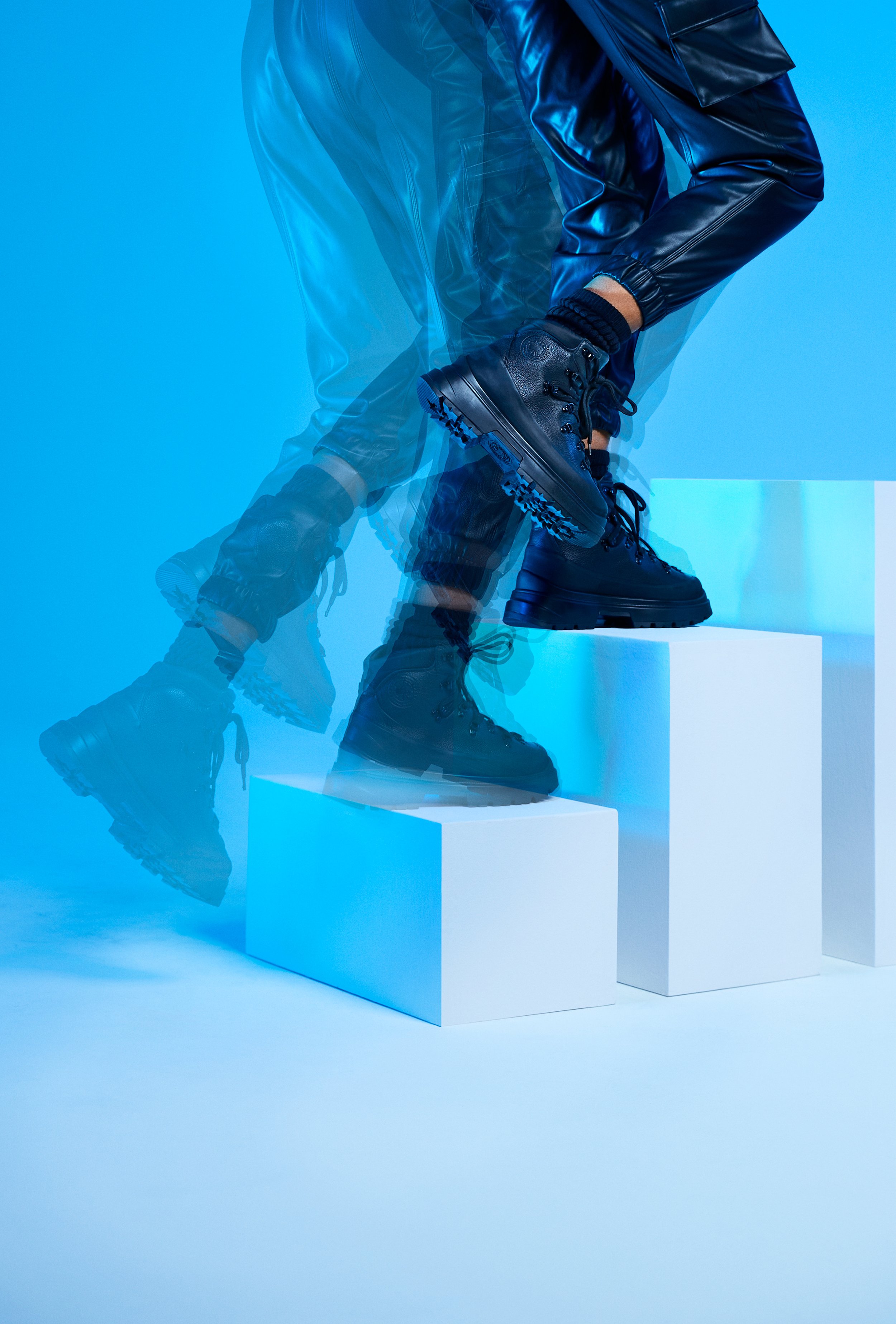 Close-up of a person in black boots and black leather pants stepping up on white platforms, with a blue background creating a dynamic motion effect.