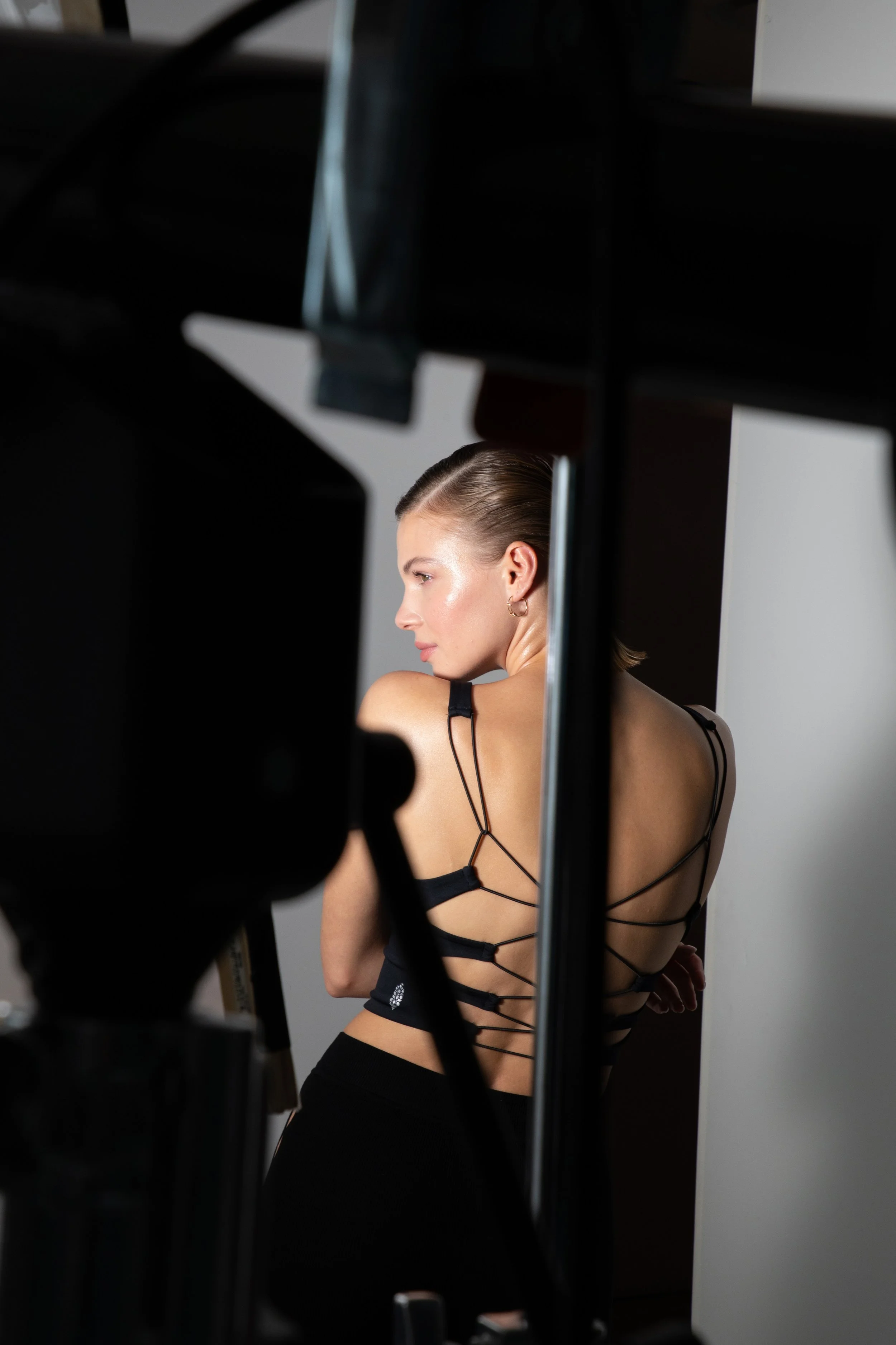 A woman with slicked-back hair and earrings wearing a black strappy backless top posing in front of a white background, seen through camera equipment.