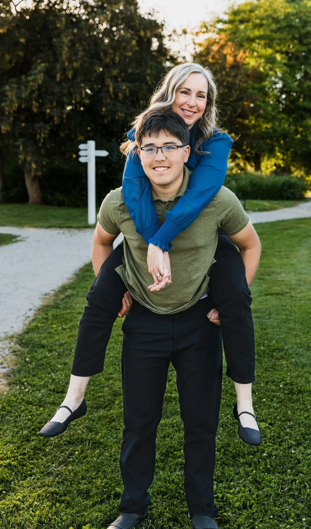 A woman with long wavy hair wearing a blue dress shirt and black shoes is given a piggyback ride from her son with black hair, glasses, and a green polo shirt in at London Farm during family session at sunset.
