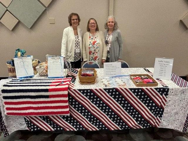 Three women standing behind a table decorated with an American flag tablecloth, showcasing baskets and signs, at a patriotic event.