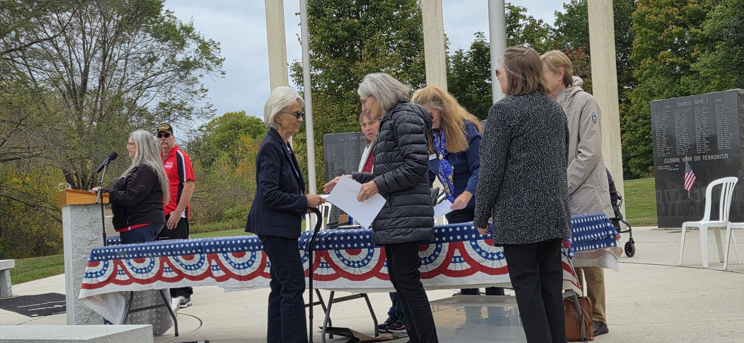 People gathered at Sheboygan Veterans Memorial around a table with patriotic decorations, participating in a ceremony, with a woman speaking at a podium and others exchanging documents.