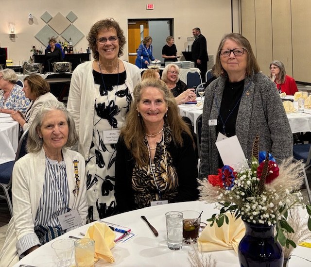 Four women posing for a photo at a formal event, with tables, chairs, and other guests in the background.