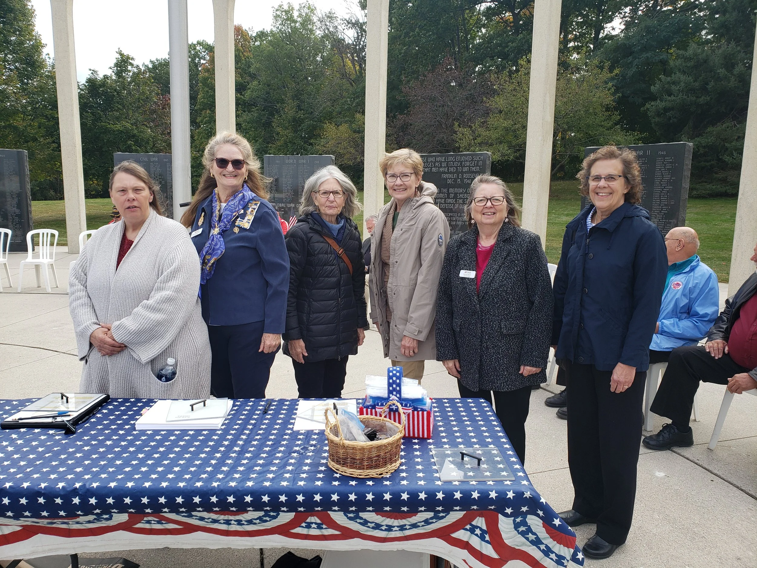 Six women standing in front of a memorial at an outdoor event, with a table decorated with an American flag-themed cloth, small American flags, and a basket in the foreground.