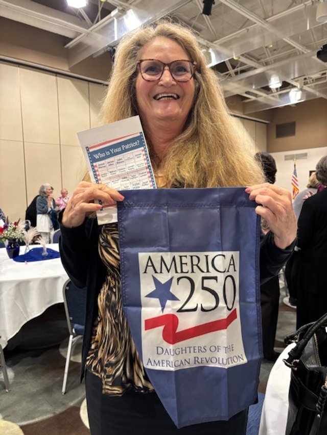 A smiling woman with glasses and long blonde hair holding a blue flag with America 250, Daughters of the American Revolution, and American Revolution symbols, in a banquet hall.
