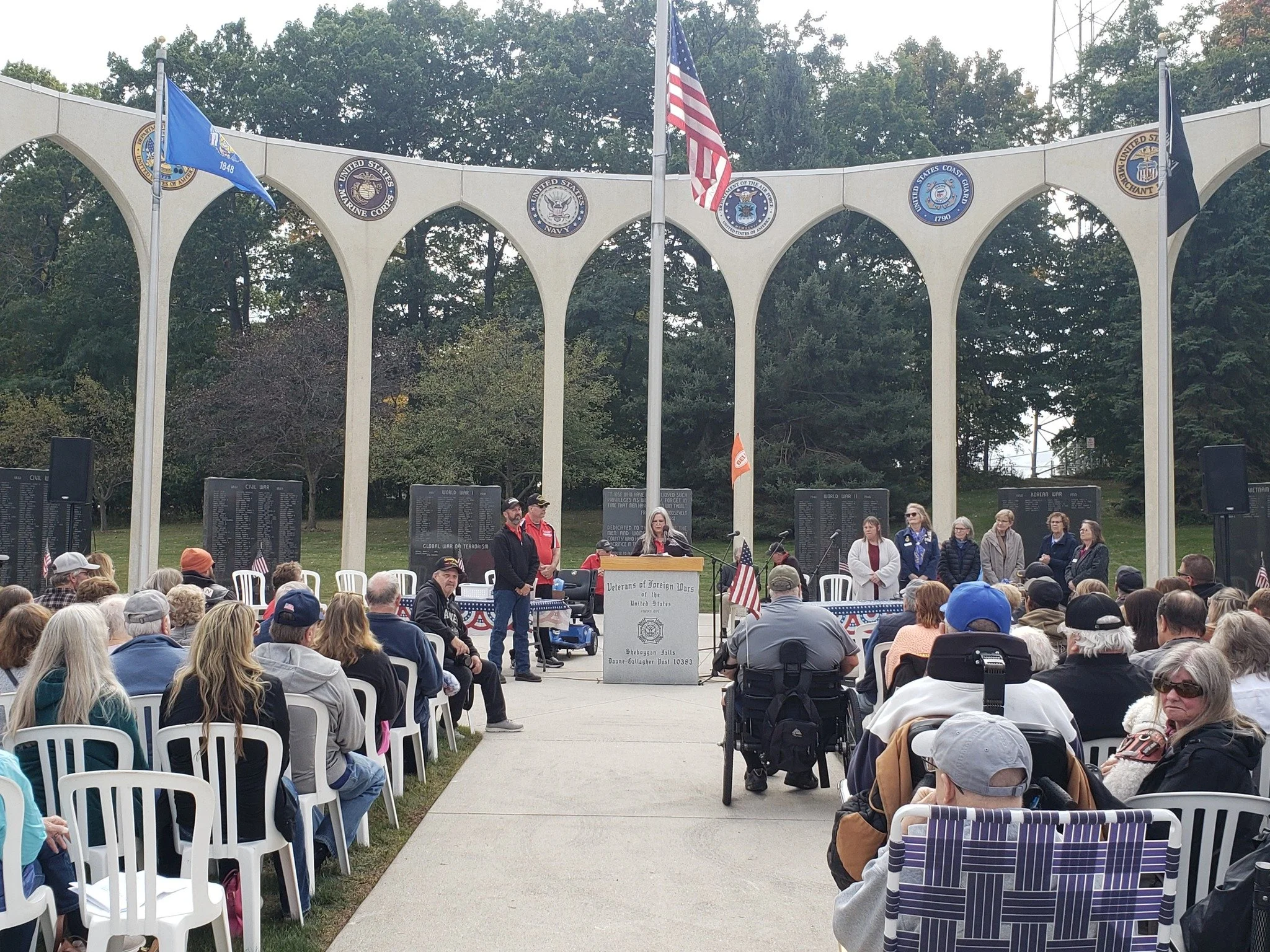 A gathering at a Wisconsin Veterans of Foreign Wars memorial ceremony with people seated and standing, American flags, and commemorative plaques in the background.