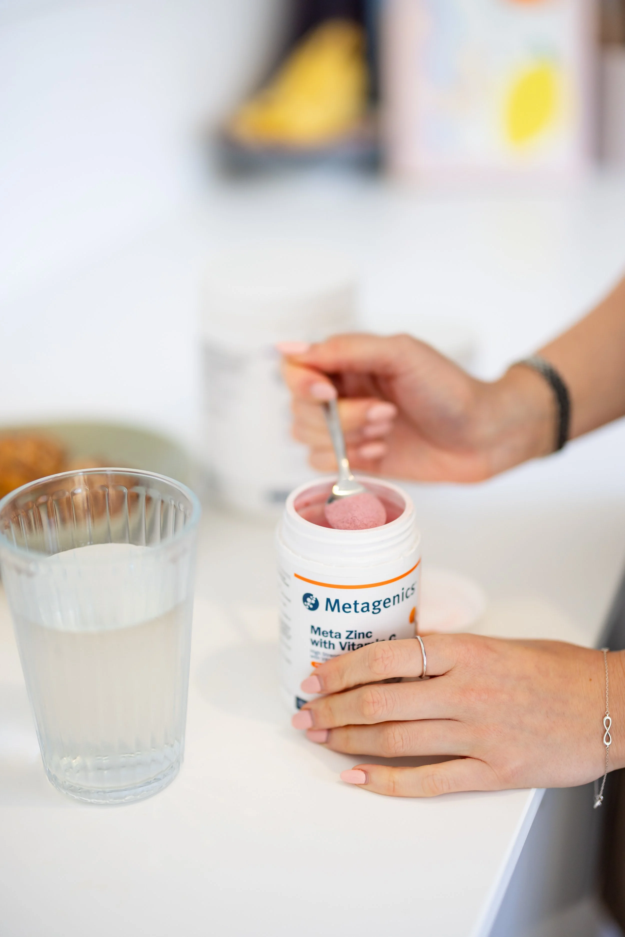 A person opening a white bottle of dietary supplement labeled 'Metagenics Meta Zinc with Vitamin C' with their right hand, on a white table with a transparent glass of water and some food items in the background.
