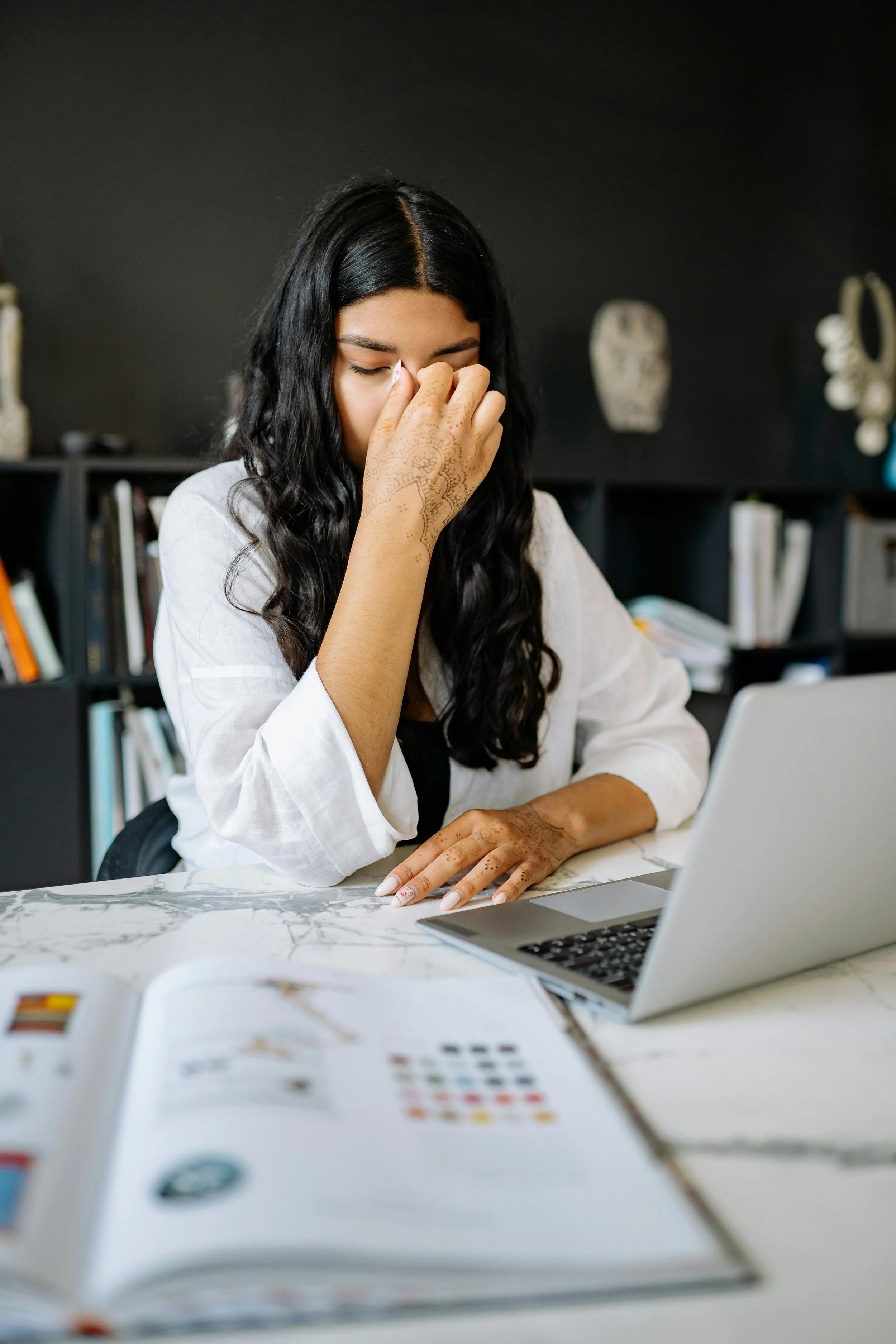 A woman with long black hair and henna on her hands, sitting at a marble table with a laptop and an open magazine, appears stressed or overwhelmed.
