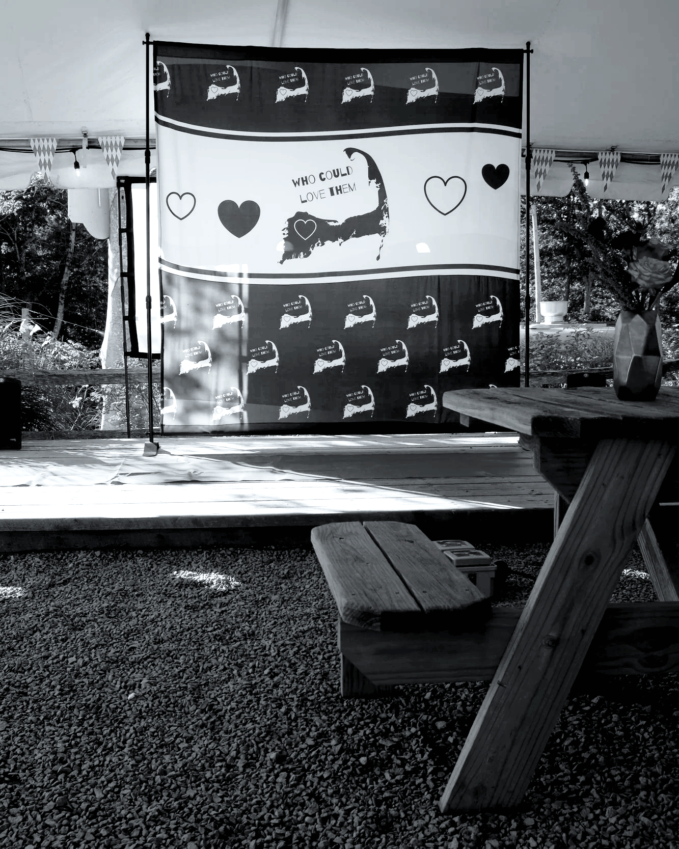 A stage with a backdrop featuring a map of cape cod and the words "Who could love them" surrounded by heart shapes, set up outdoors with picnic tables nearby.