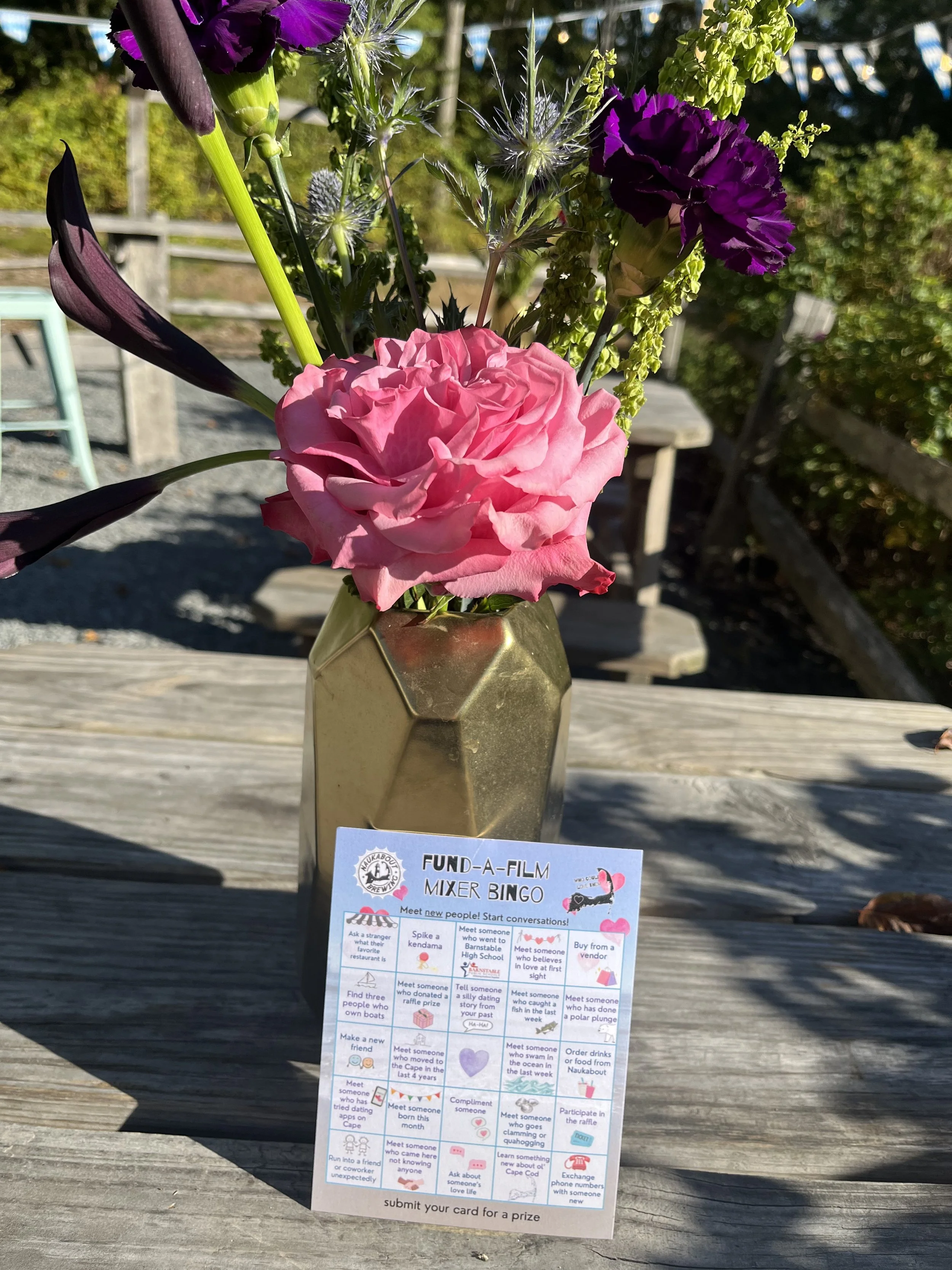 A bouquet of purple, pink, and green flowers in a gold geometric vase on a wooden surface outdoors, with a bingo card titled 'Fund-A-Film Mixer Bingo' placed in front of the vase.