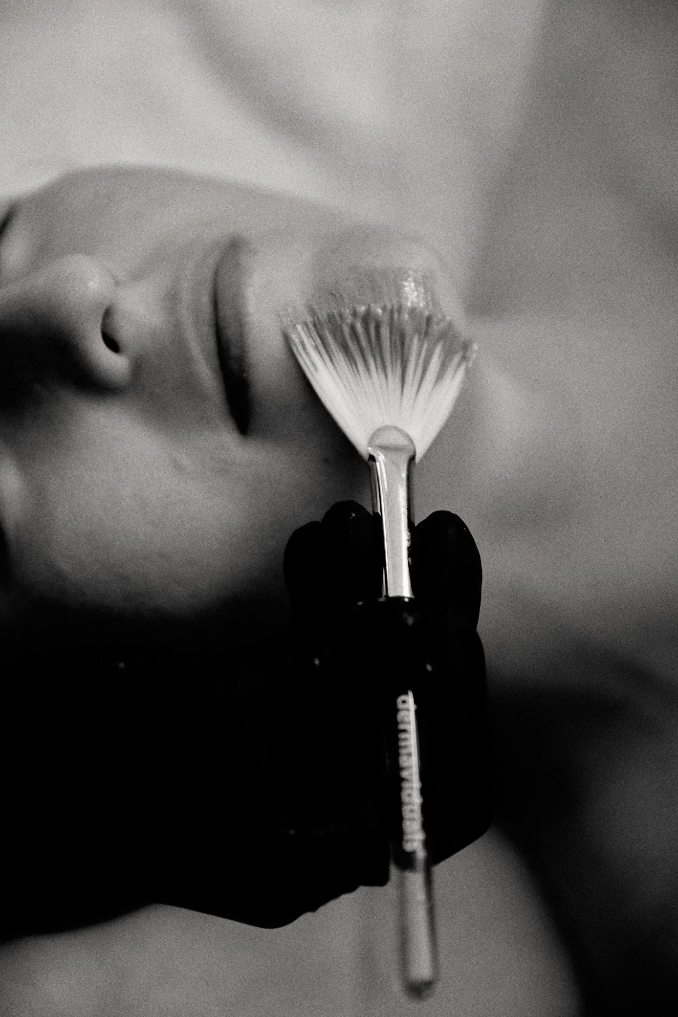 Close-up of a cosmetic brush held by a gloved hand applying product to a person's face.