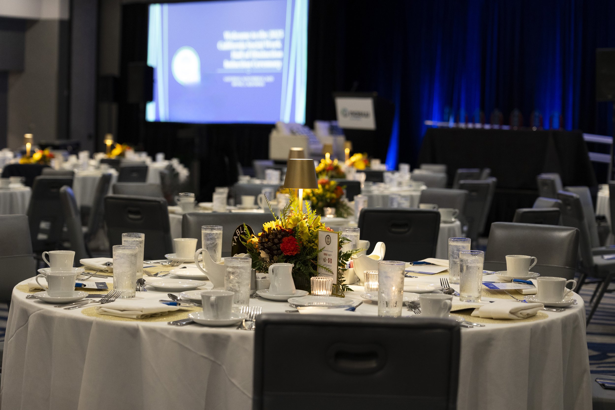 A banquet table set for a formal event with white tablecloths, floral centerpieces with yellow and red flowers, and place settings including cups, plates, silverware, and water glasses. In the background, a stage with a large screen and dark curtains