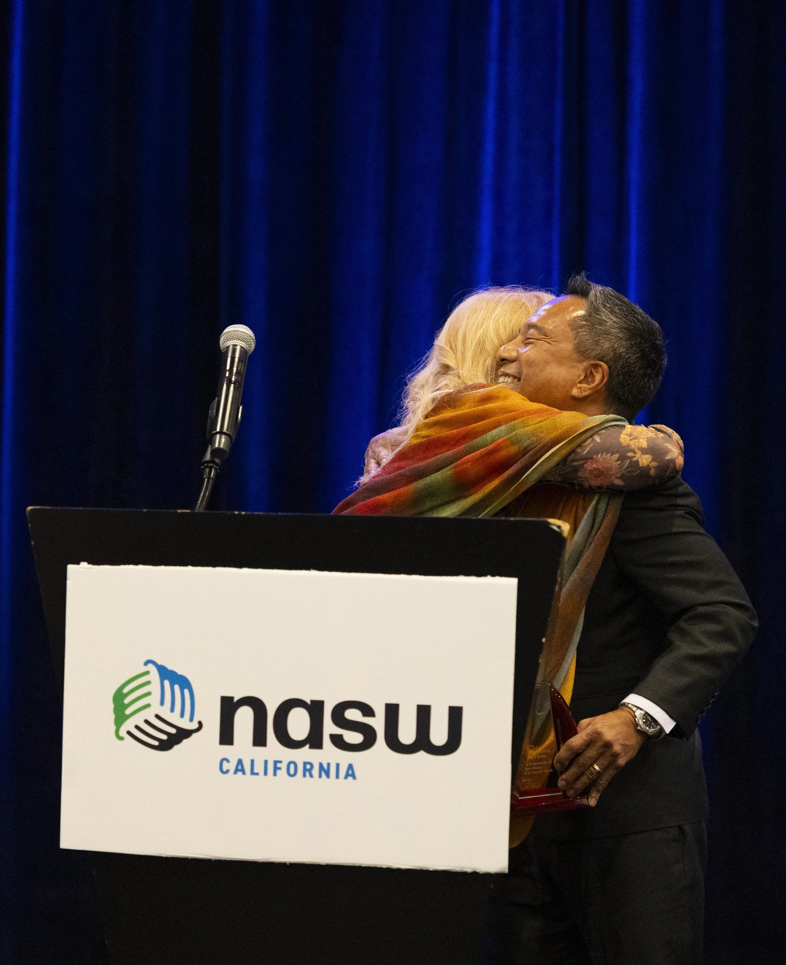 Two people hugging on stage at a NASW California event, with a podium and microphone in front of them, and dark blue curtains in the background.