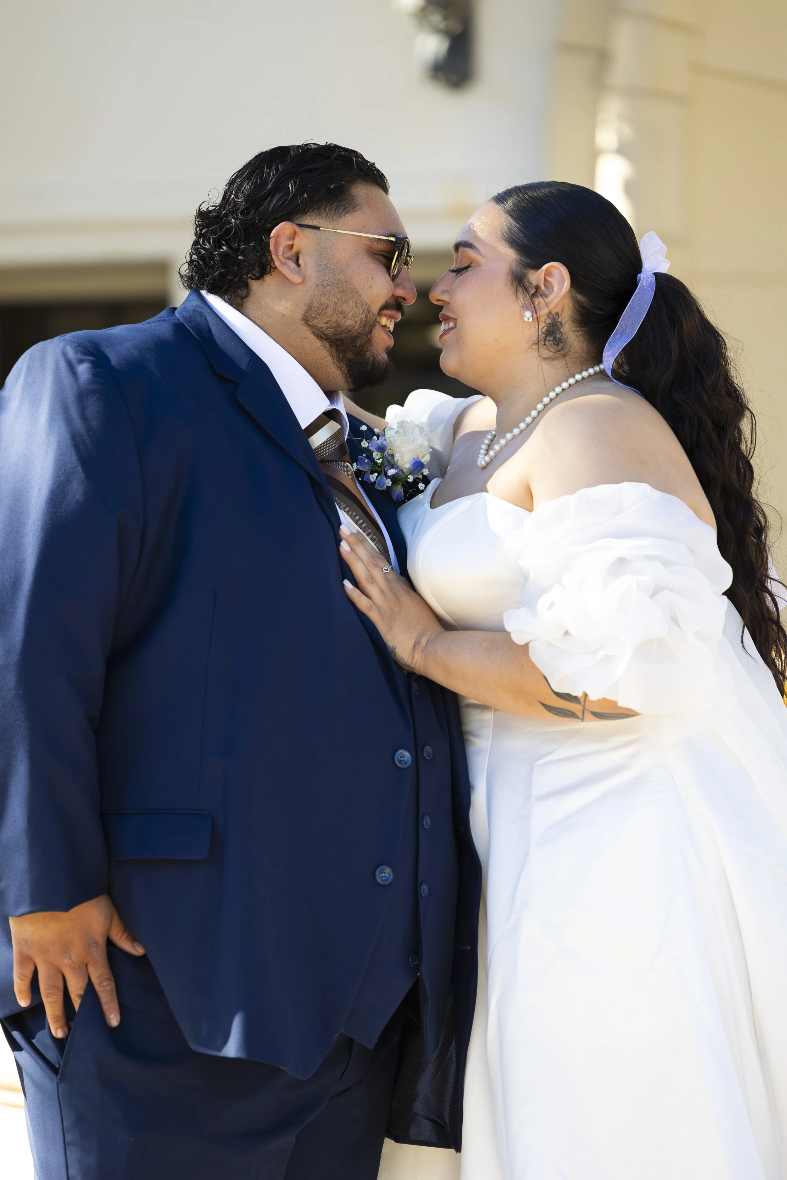 A happy couple dressed in wedding attire, facing each other closely with their foreheads touching, outdoors with a building in the background.