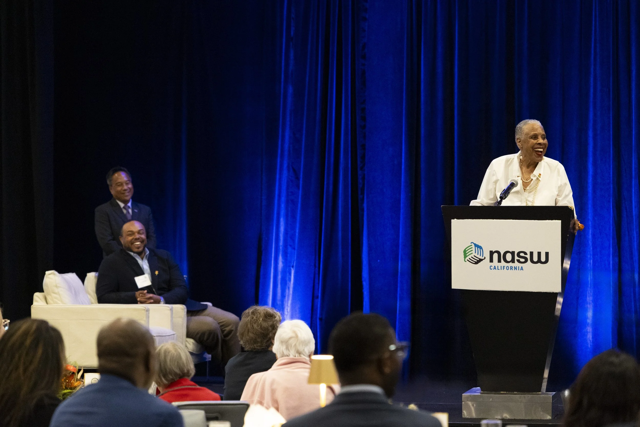 An elderly woman stands at a podium speaking at a conference with a blue curtain background and an audience watching, while two men sit on a couch to the side.