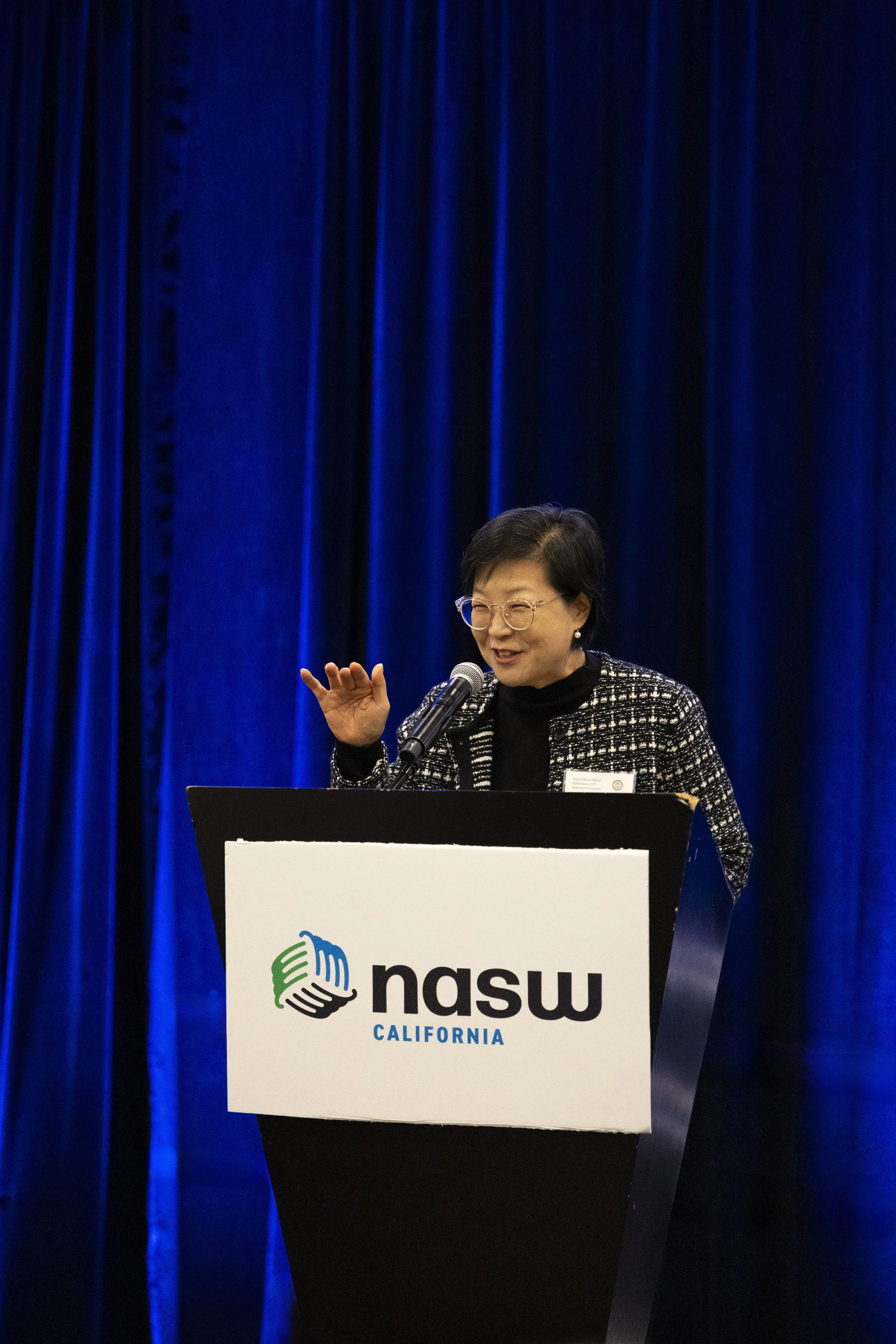 Woman speaking at a NASW California event, standing behind a podium with NASW logo and blue curtains in background.