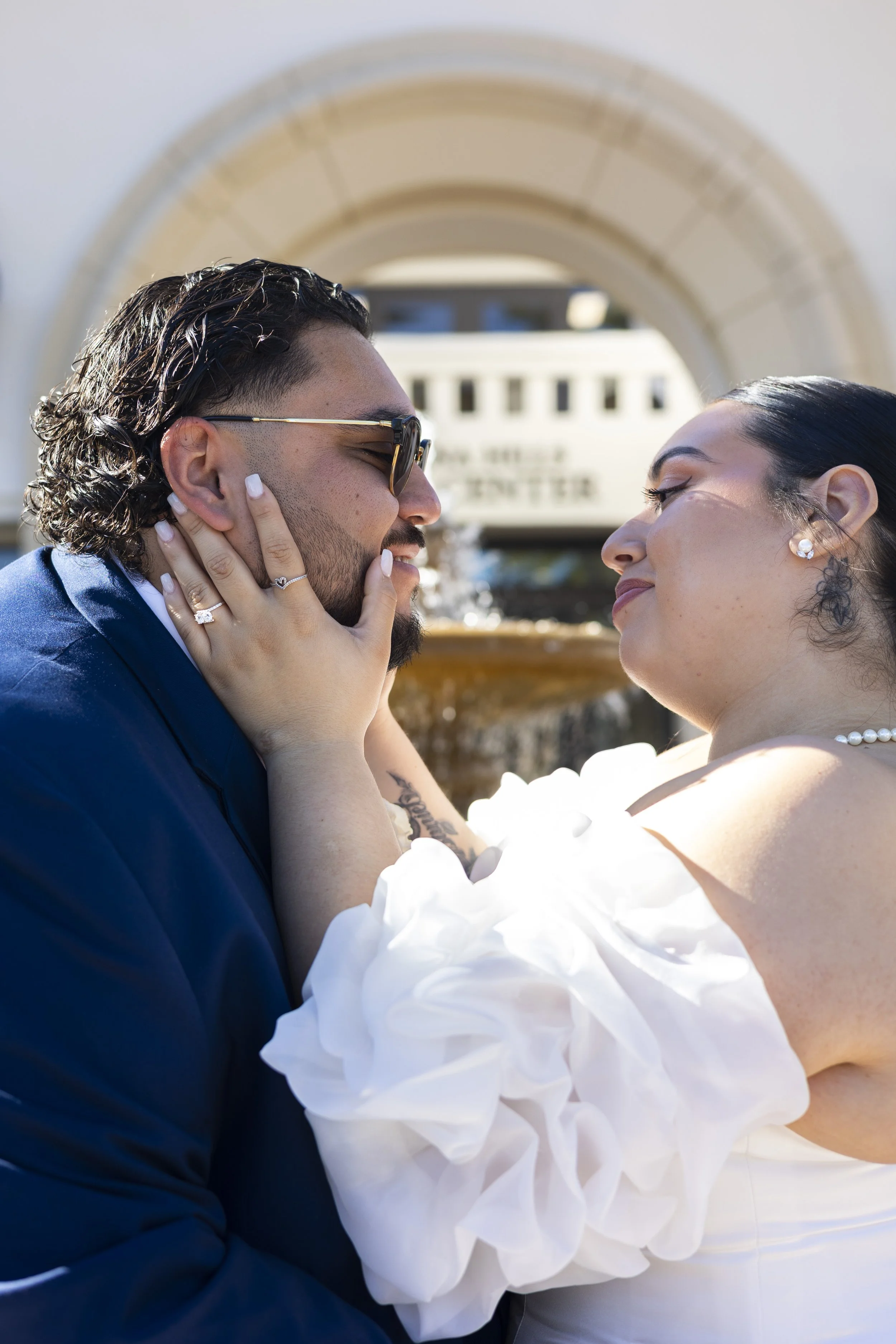 A couple during their wedding or engagement photos, close-up, with a fountain and a building in the background, sharing an intimate moment.