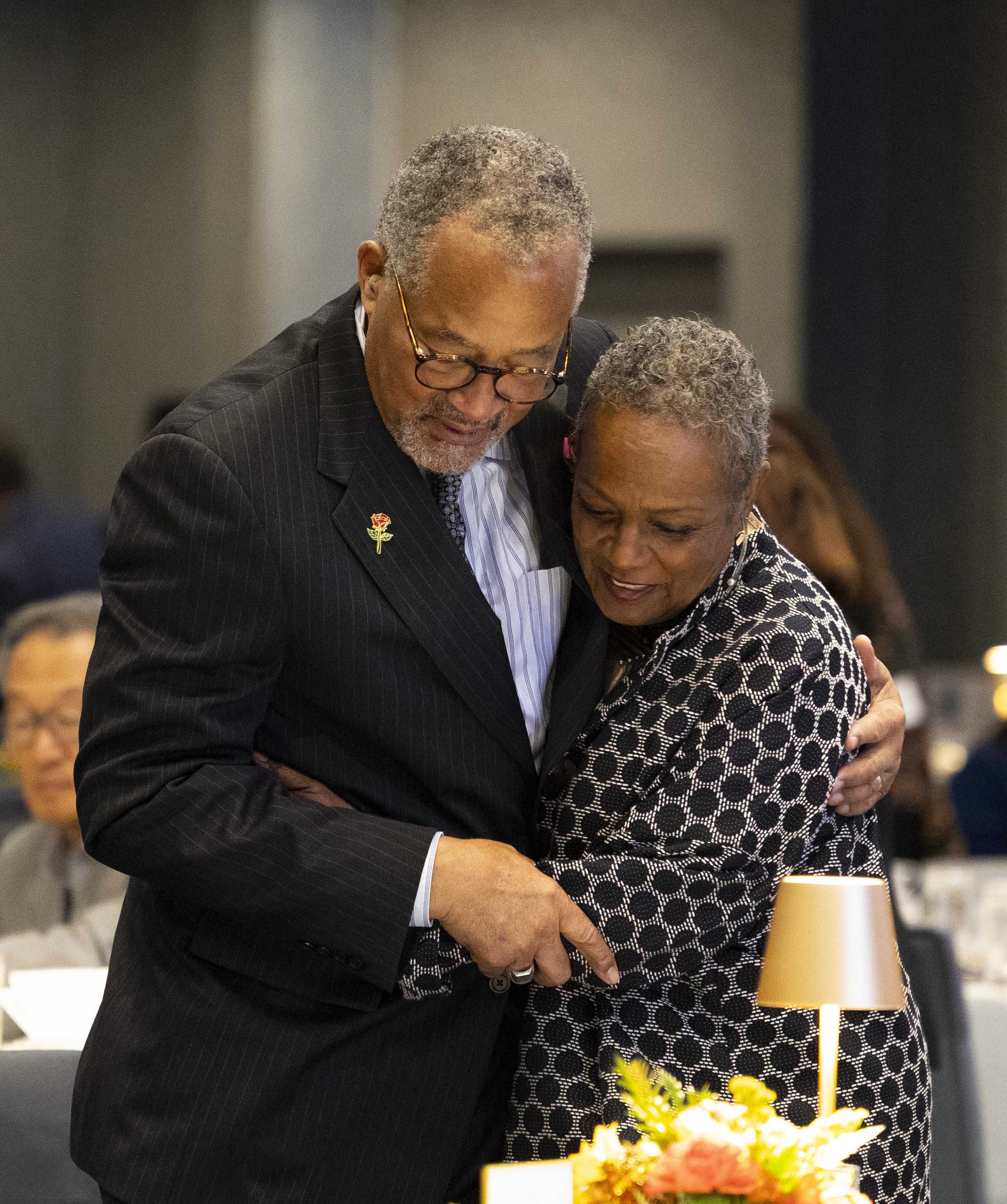 An elderly man and woman embrace each other affectionately at an event, with the man wearing a pin on his suit lapel and the woman in a polka dot blouse, surrounded by a warmly lit setting with a floral arrangement and other guests in the background.