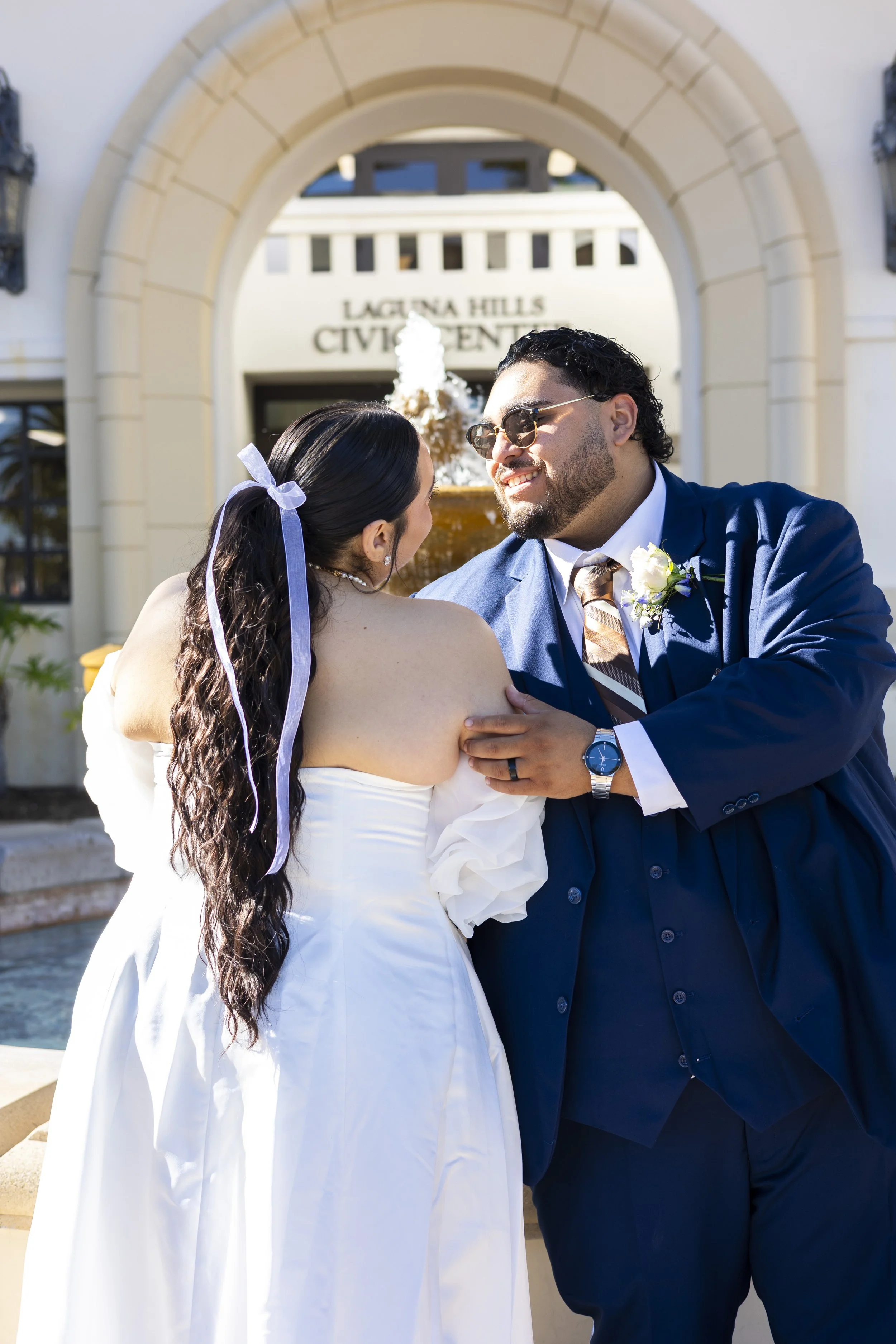 A newlywed couple sharing a moment in front of Laguna Hills Civic Center, with the groom smiling and the bride facing away, wearing a white wedding dress and a lavender ribbon in her long, wavy hair.