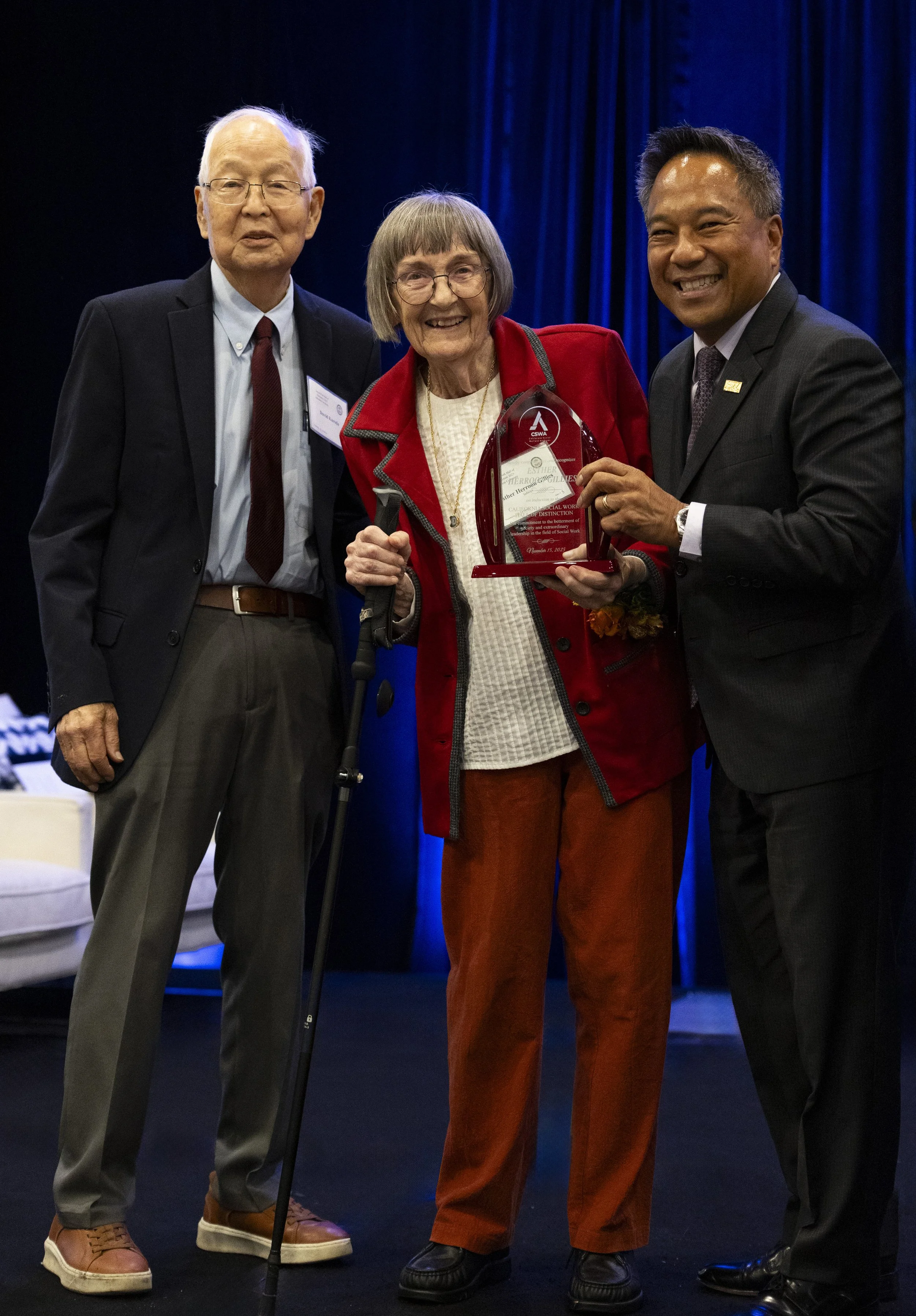 Three people on stage at an awards ceremony, one woman holding a trophy, flanked by two men in suits.