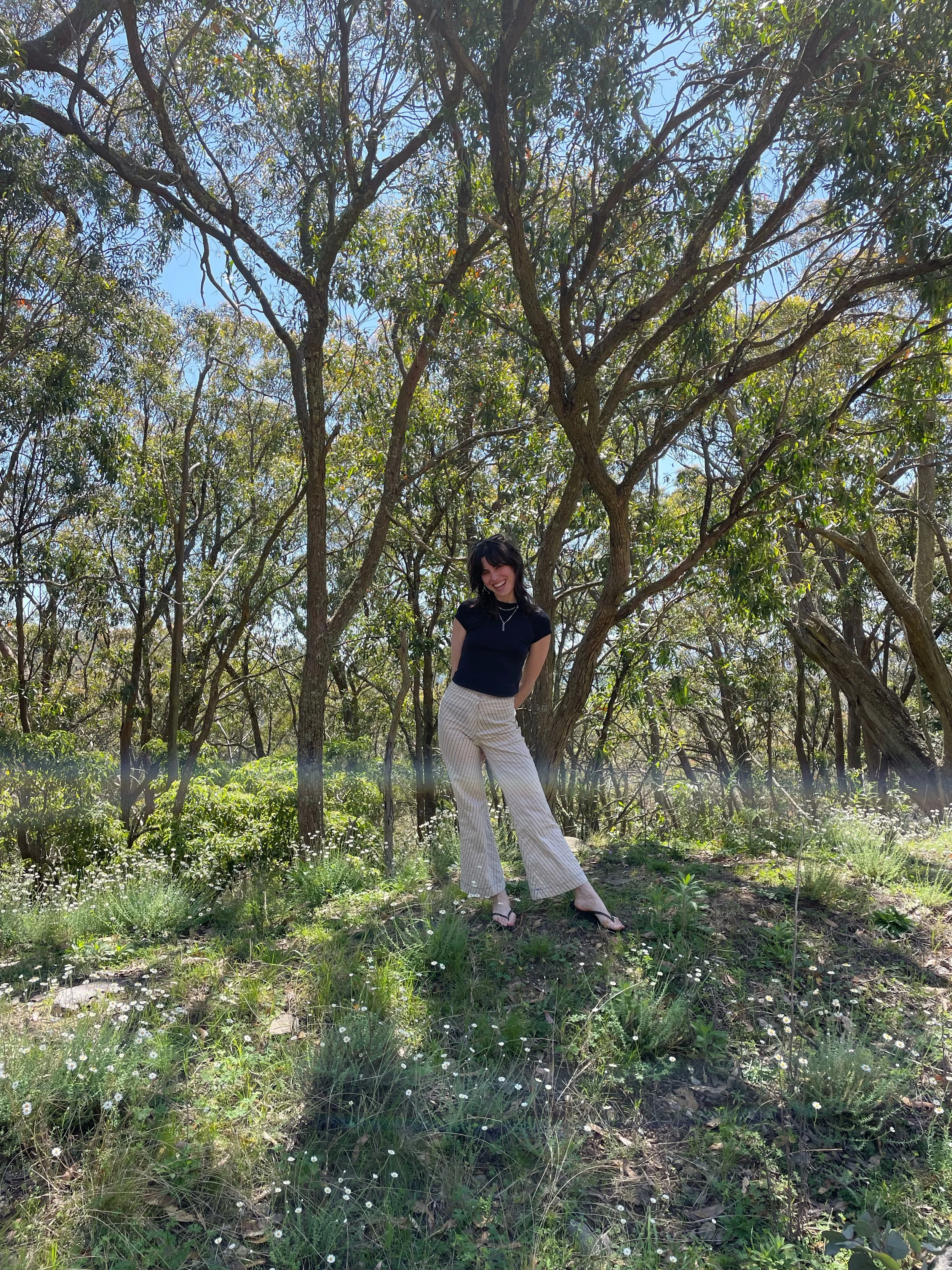 A woman standing in a wooded outdoor area with trees, grass, and small white flowers, smiling at the camera.