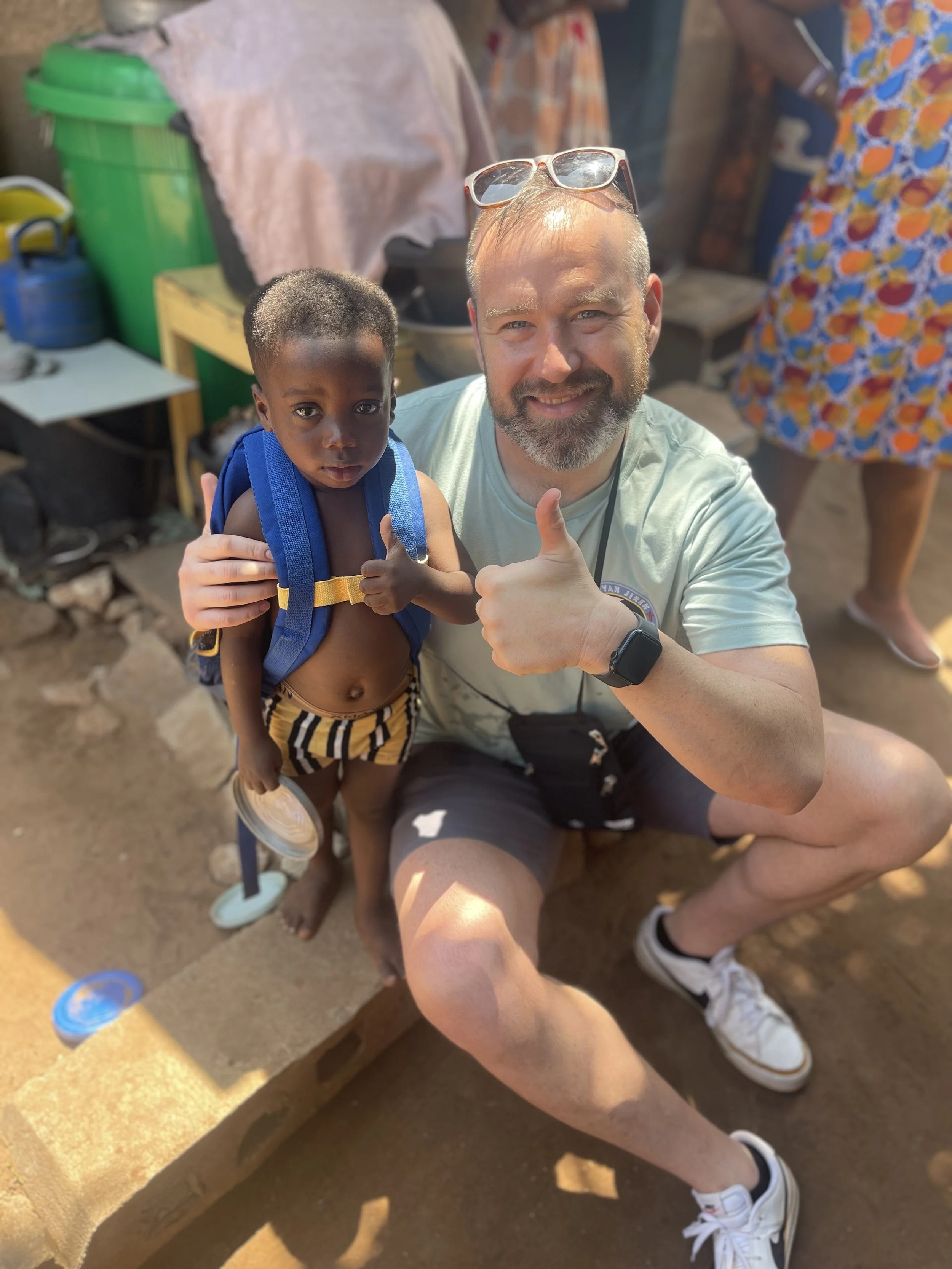 A man with a beard, wearing sunglasses on his head, is squatting down and giving a thumbs-up. He is holding a young boy with dark skin, short hair, and a blue backpack. The boy is shirtless, wearing striped shorts, and holding a spoon. They are outdoors with dirt ground and various supplies and people in the background.