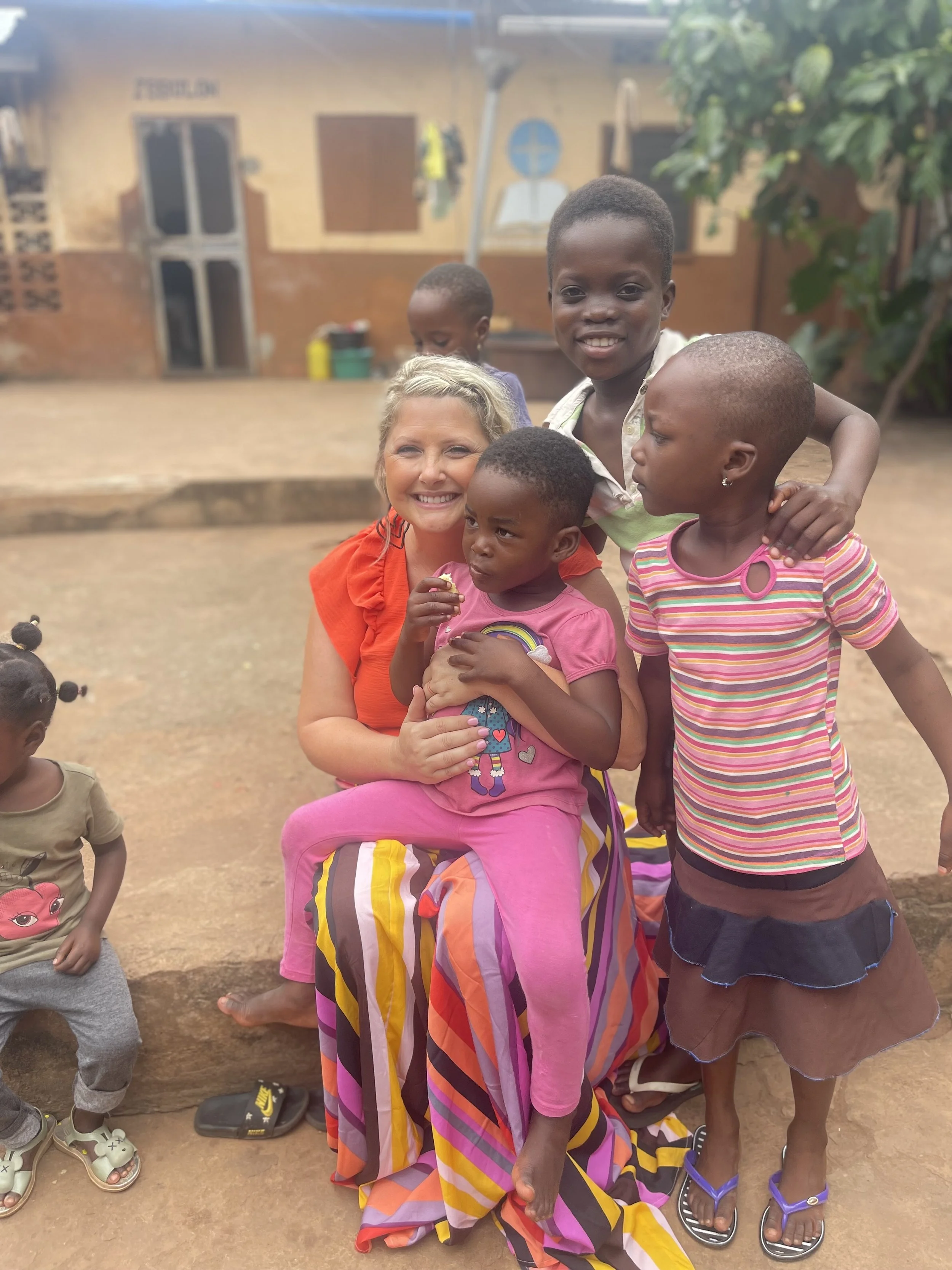 A woman with blonde hair smiles while sitting on a stone step, surrounded by four children, outside a building with painted walls and a few plants. The children are interacting with her, and one is sitting on her lap.