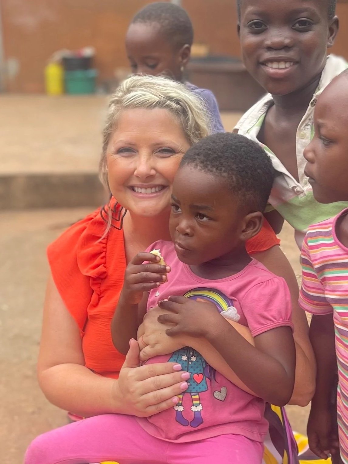 A smiling woman hugging a young girl in pink, surrounded by three other children, in an outdoor setting.