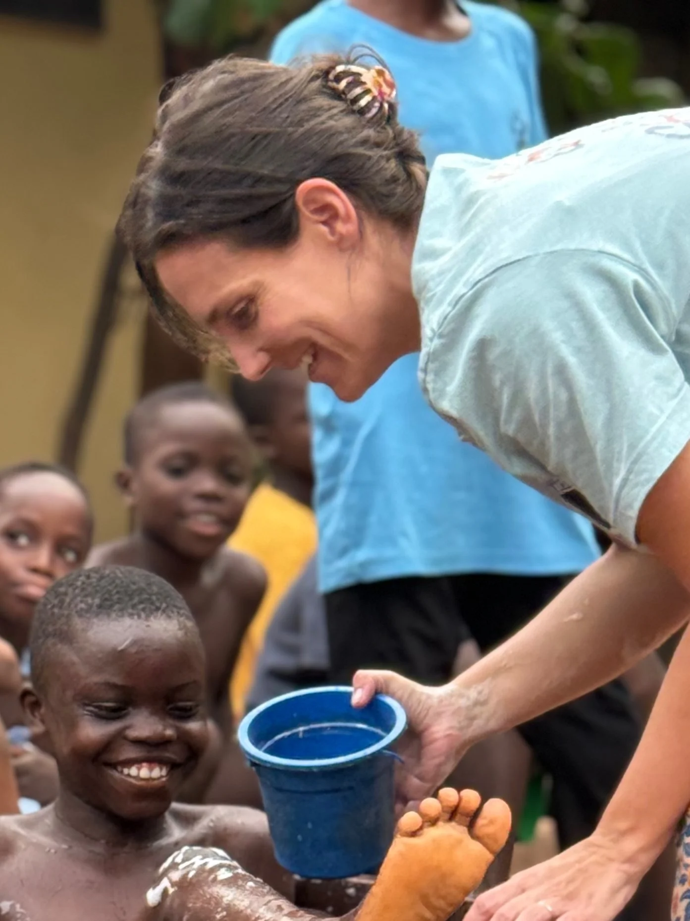 A woman giving a foot bath to a smiling young boy surrounded by other children outdoors.