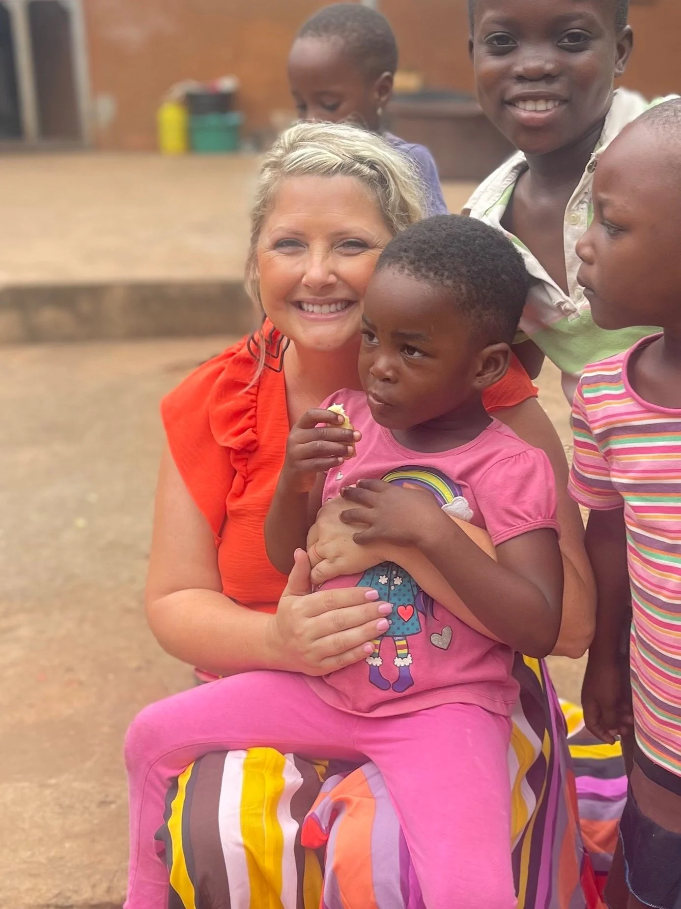A smiling woman hugging a young girl in pink, surrounded by three other children, in an outdoor setting.
