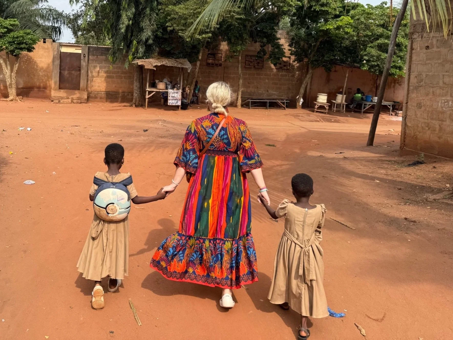 A woman with gray hair wearing a colorful, patterned dress walking on a dirt road in an African village, holding hands with two young girls in beige dresses, one wearing a backpack, in a rural setting with trees, simple houses, and a small shop in the background.