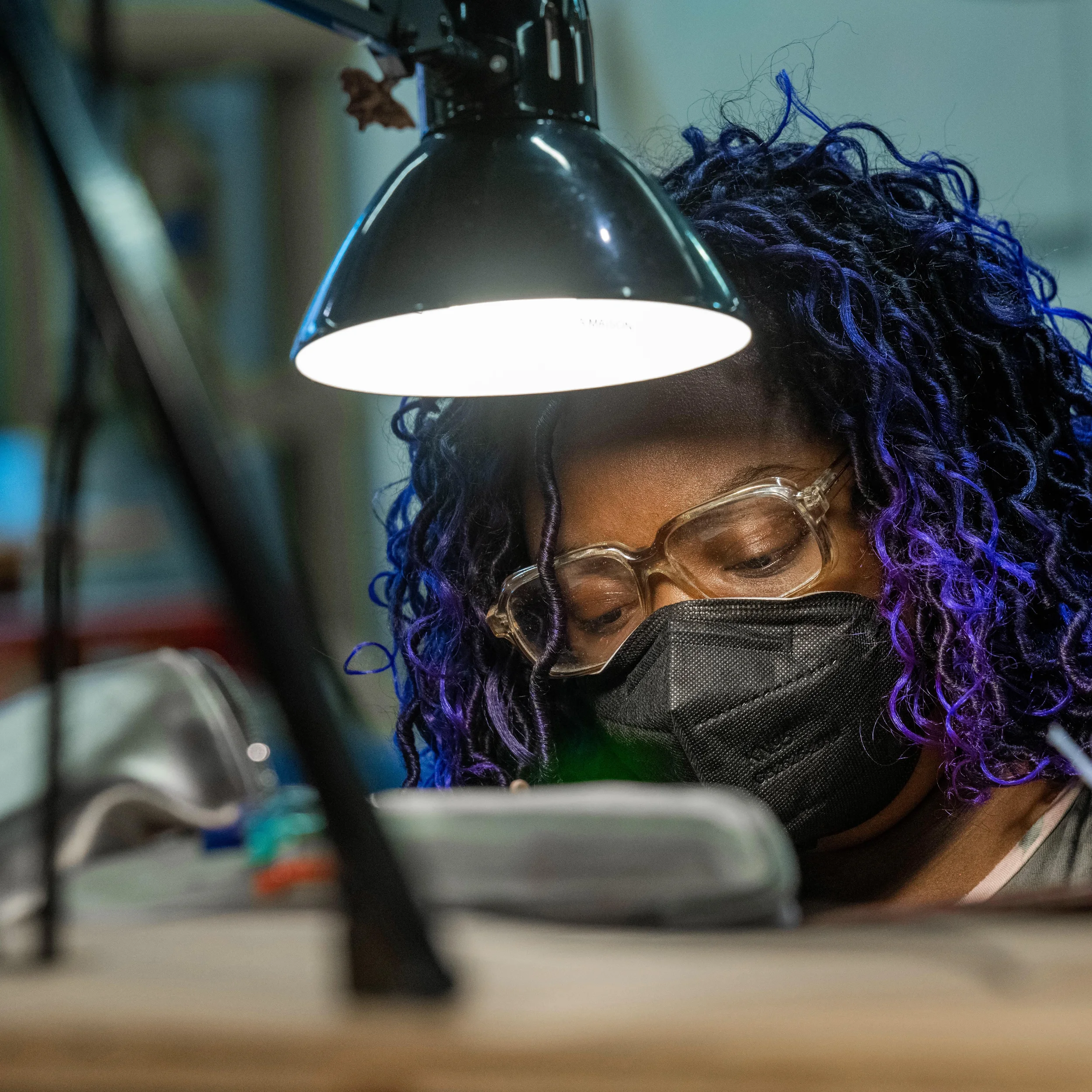 Black person with glasses, a black mask, and blue hair working under the light of a lamp