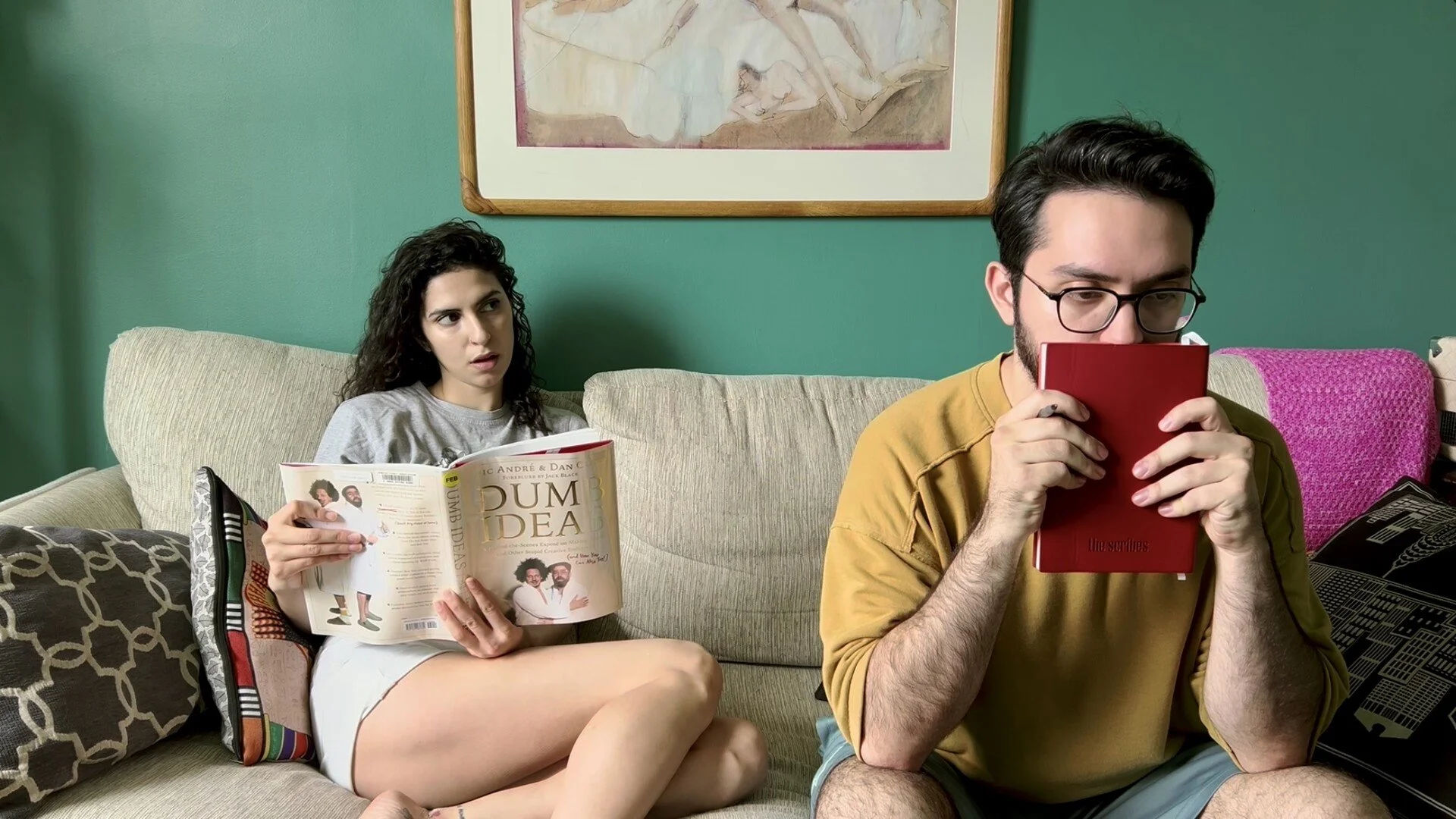 A woman and a man sit on a beige couch in a living room. The woman, with curly dark hair, is reading a magazine and looks surprised. The man, with dark hair, glasses, and a beard, is holding a small red notebook and looking at it while sitting cross-legged. The background features a green wall, a framed painting, and colorful pillows.