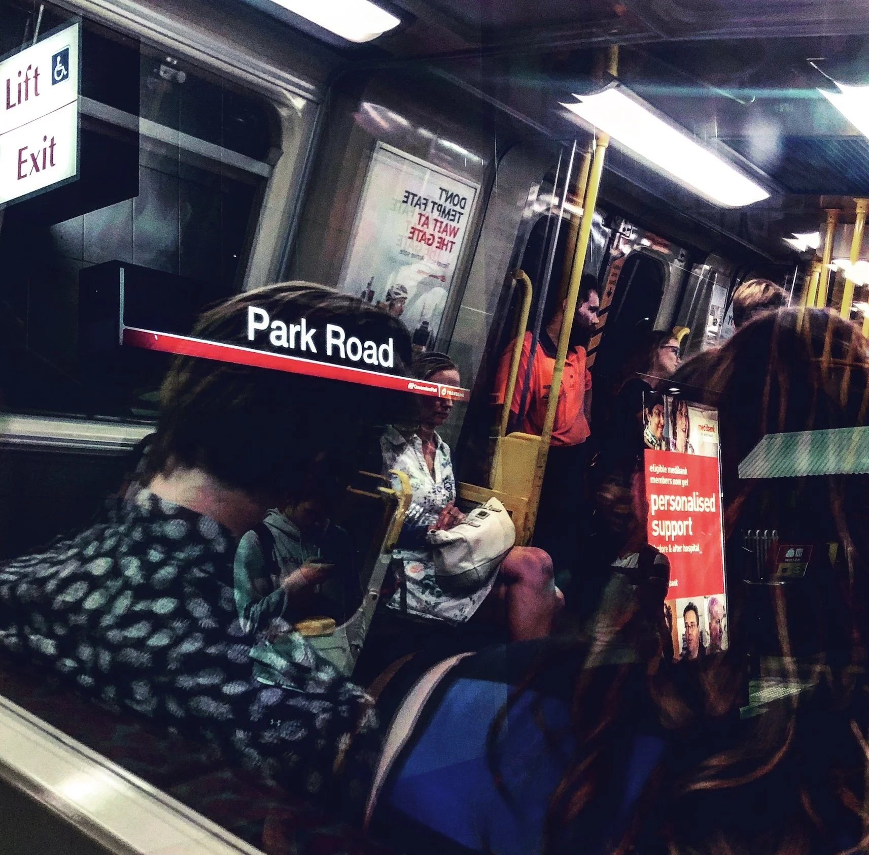 This is a photo taken by Jennine Heymer. View of a train interior with passengers seated and standing, a window reflects the scene outside. The train is at Park Road station, indicated by the station sign, and features advertisements on the walls.