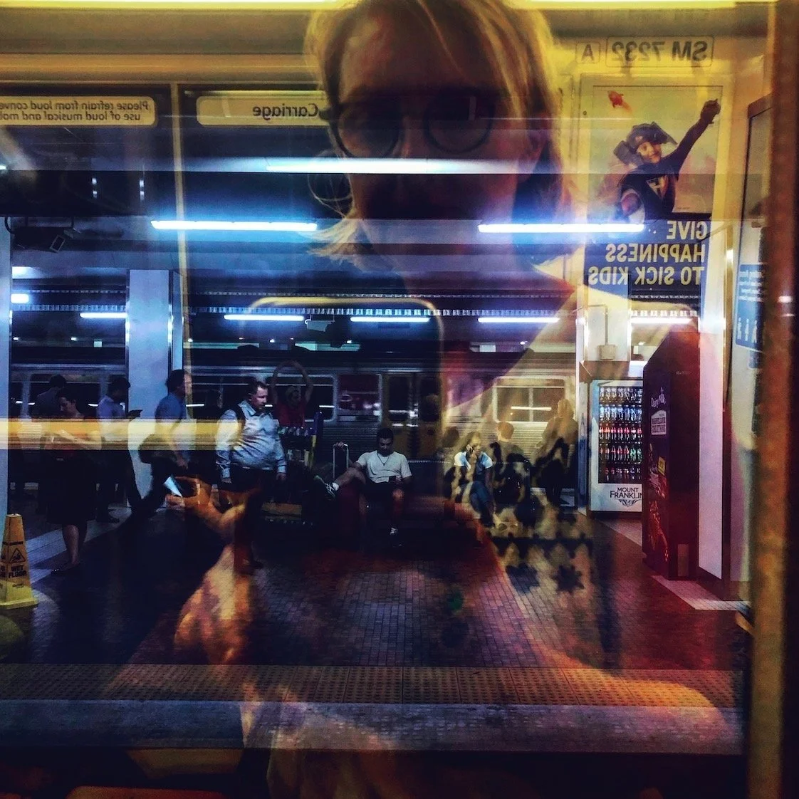 This is a photo taken by Jennine Heymer on a train. Reflection of a woman with glasses taking a picture at a train station, with people waiting and vending machines visible behind the glass.