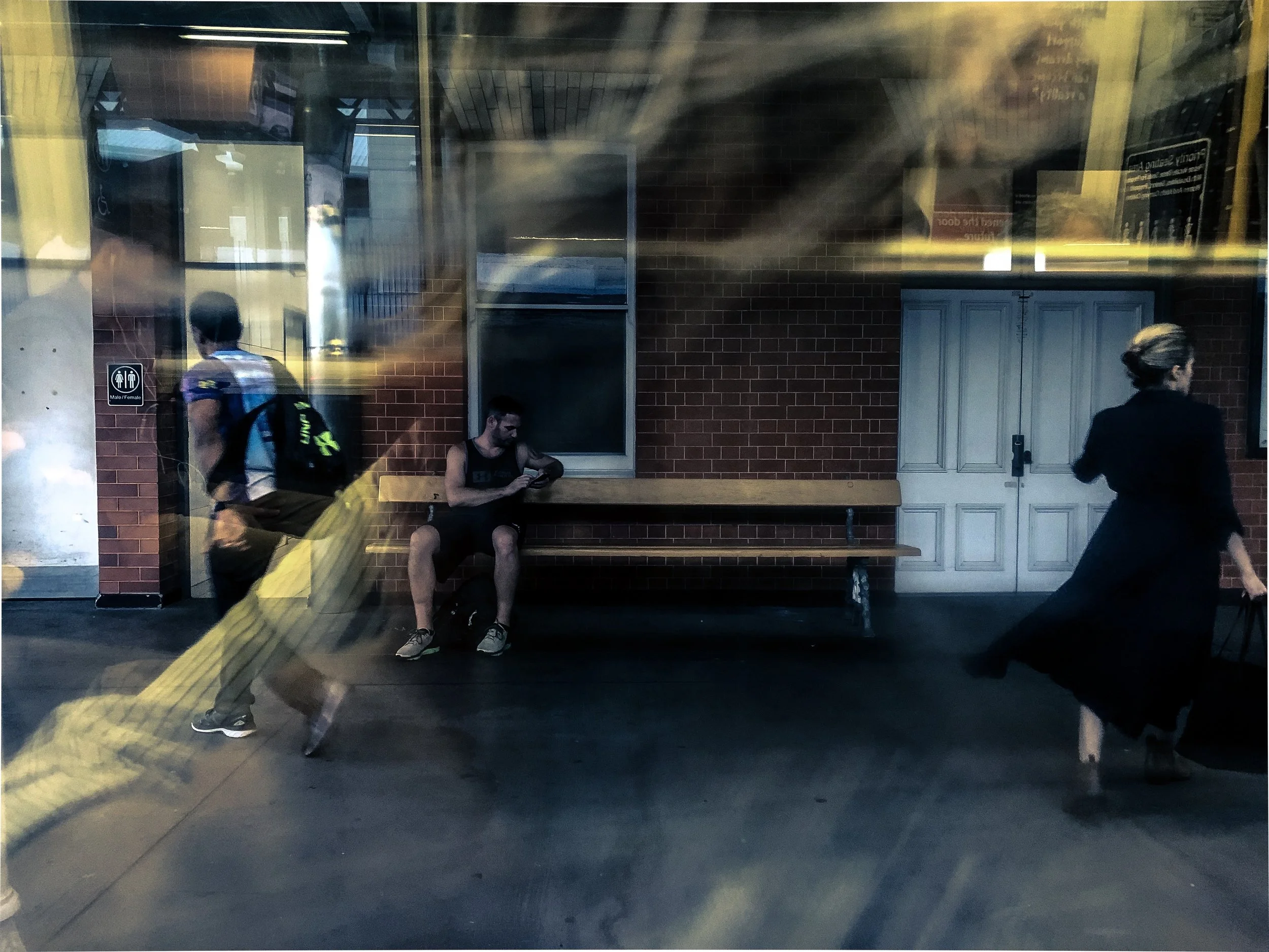 A photo taken by Jennine Heymer. Reflected image of a train station platform with three people: a man with a backpack, a seated man looking at his phone, and a woman walking with a bag. There are brick walls, a window, and bathroom signage.