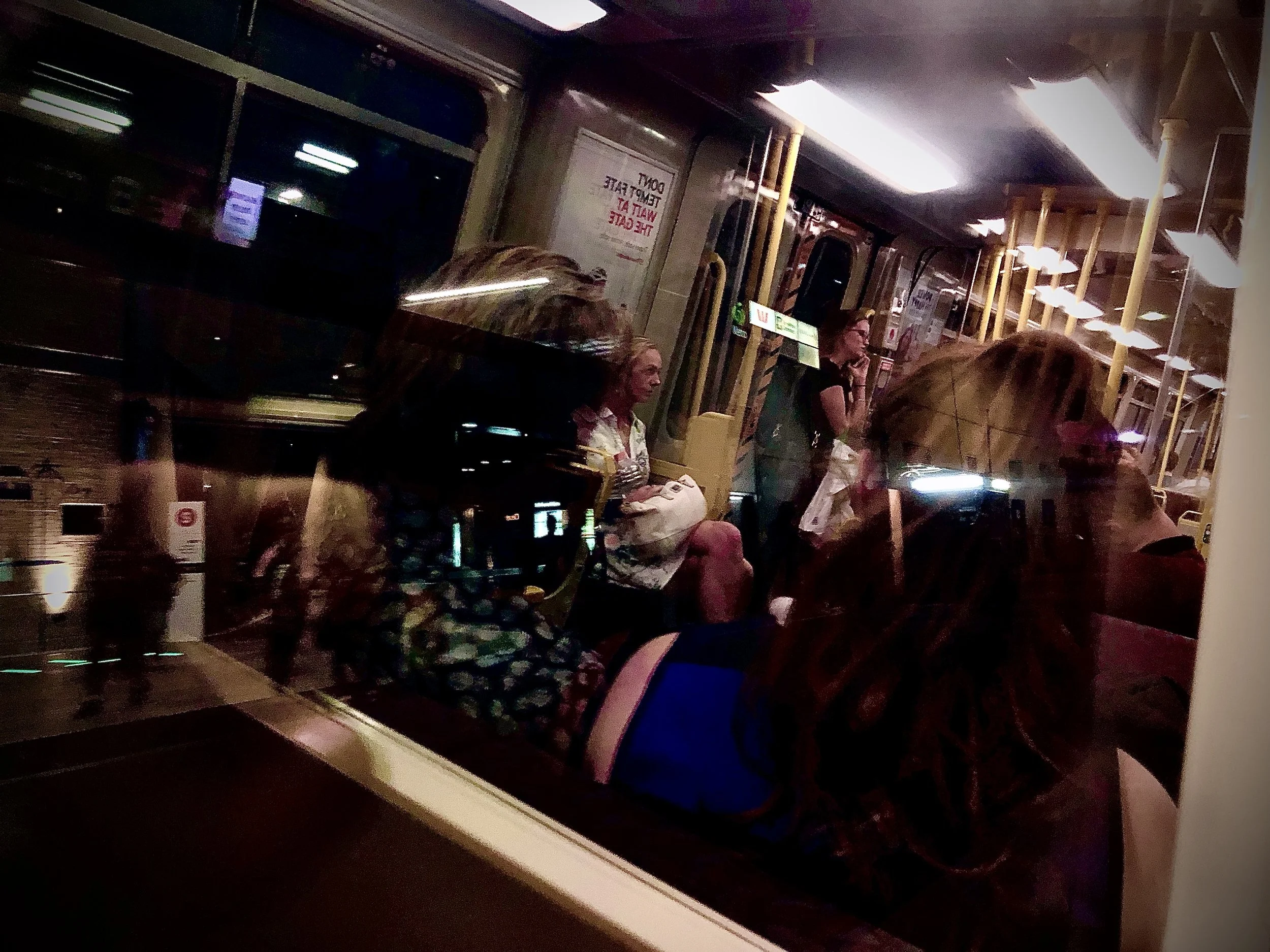 This is a photo taken by Jennine Heymer on a train. People sitting and standing inside a bus at night, seen through a window with reflections.