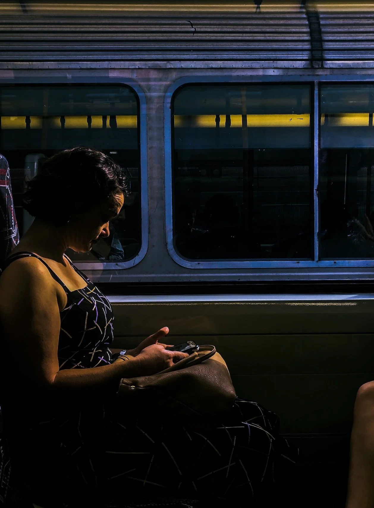 This is a photo taken by Jennine Heymer on a train. A woman with short black hair sitting on a bus, looking at her phone, with a brown purse on her lap, against the backdrop of a window showing the side of another vehicle.