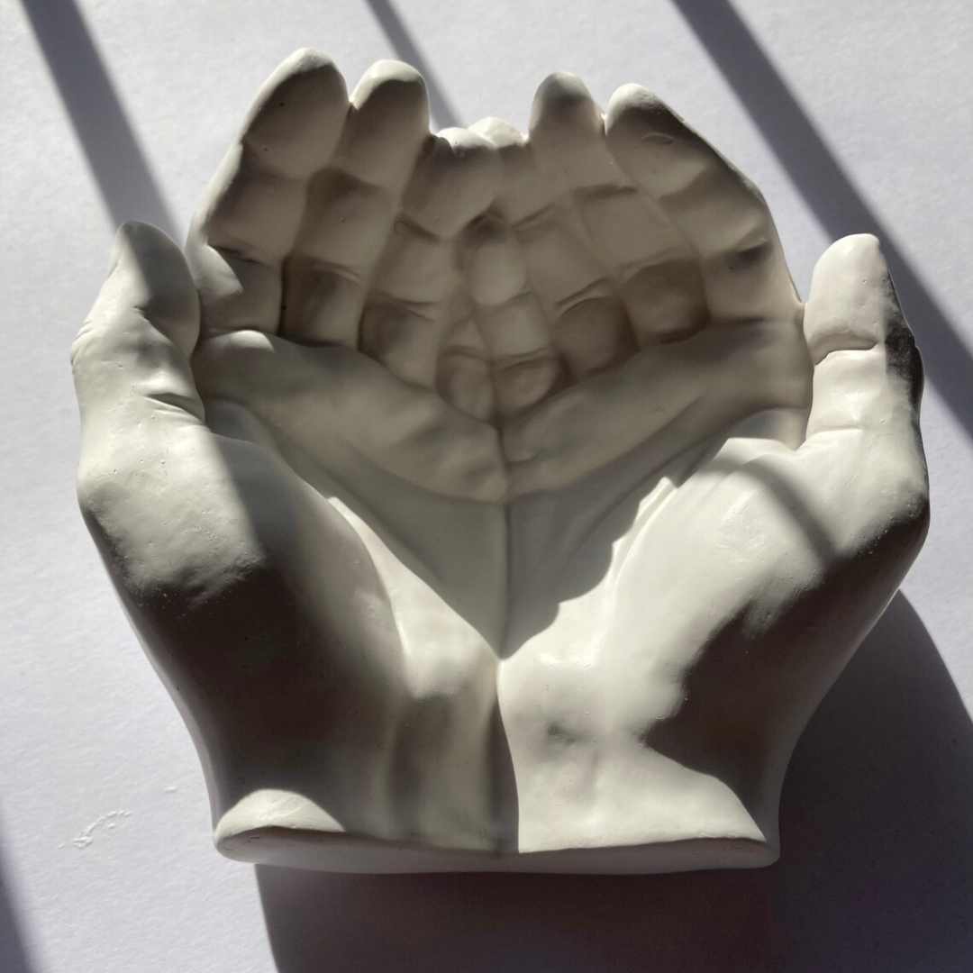 A white sculpture of a hand holding a heart with raised fingers, casting shadow on a light-colored surface.