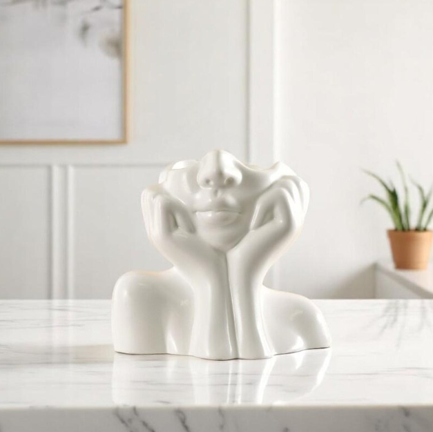 White ceramic sculpture of a face with hands on cheeks, smiling, on a marble surface.